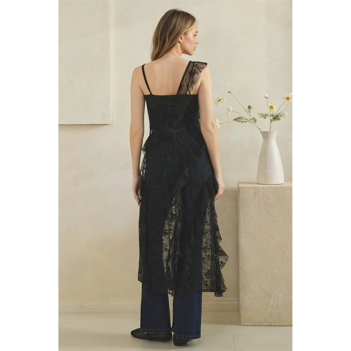 A woman stands with her back to the camera wearing a black lace and mesh asymmetrical duster over dark wide-leg jeans, in a softly lit indoor setting with a vase of flowers on a pedestal.