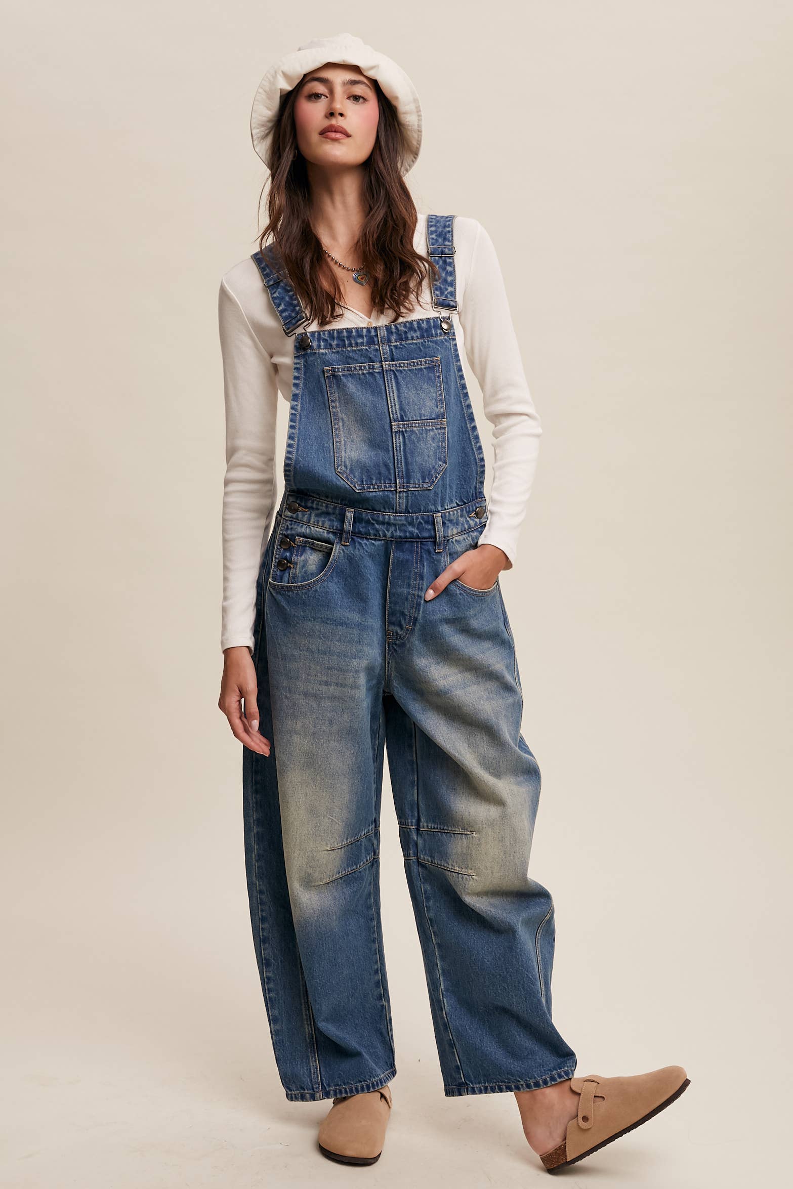 Vintage-inspired blue denim overalls with relaxed wide-leg fit, front chest pocket, and adjustable straps, styled with a white long-sleeve top and neutral clogs.