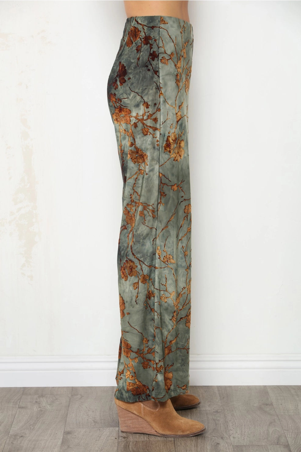 A person shown from the waist down wearing wide-leg pants with an abstract floral pattern in muted green, brown, and rust tones, paired with tan wedge ankle boots, standing against a white wall and wood floor.