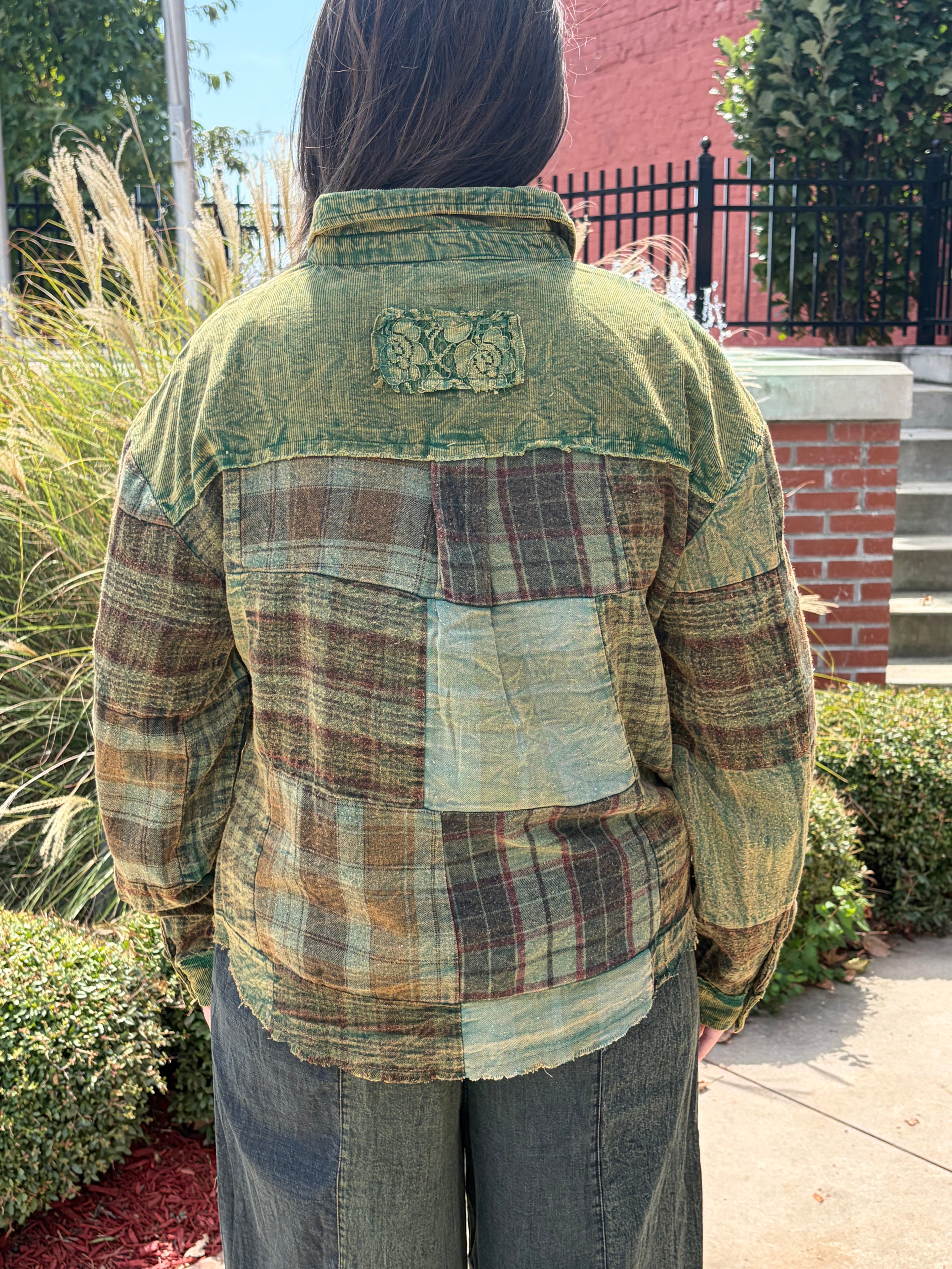 A person viewed from behind wearing a vintage-style patchwork flannel shirt with a distressed green collar and back, brown and beige plaid body, and a large light green pocket on the back, standing outdoors in front of a brick wall and black fence.