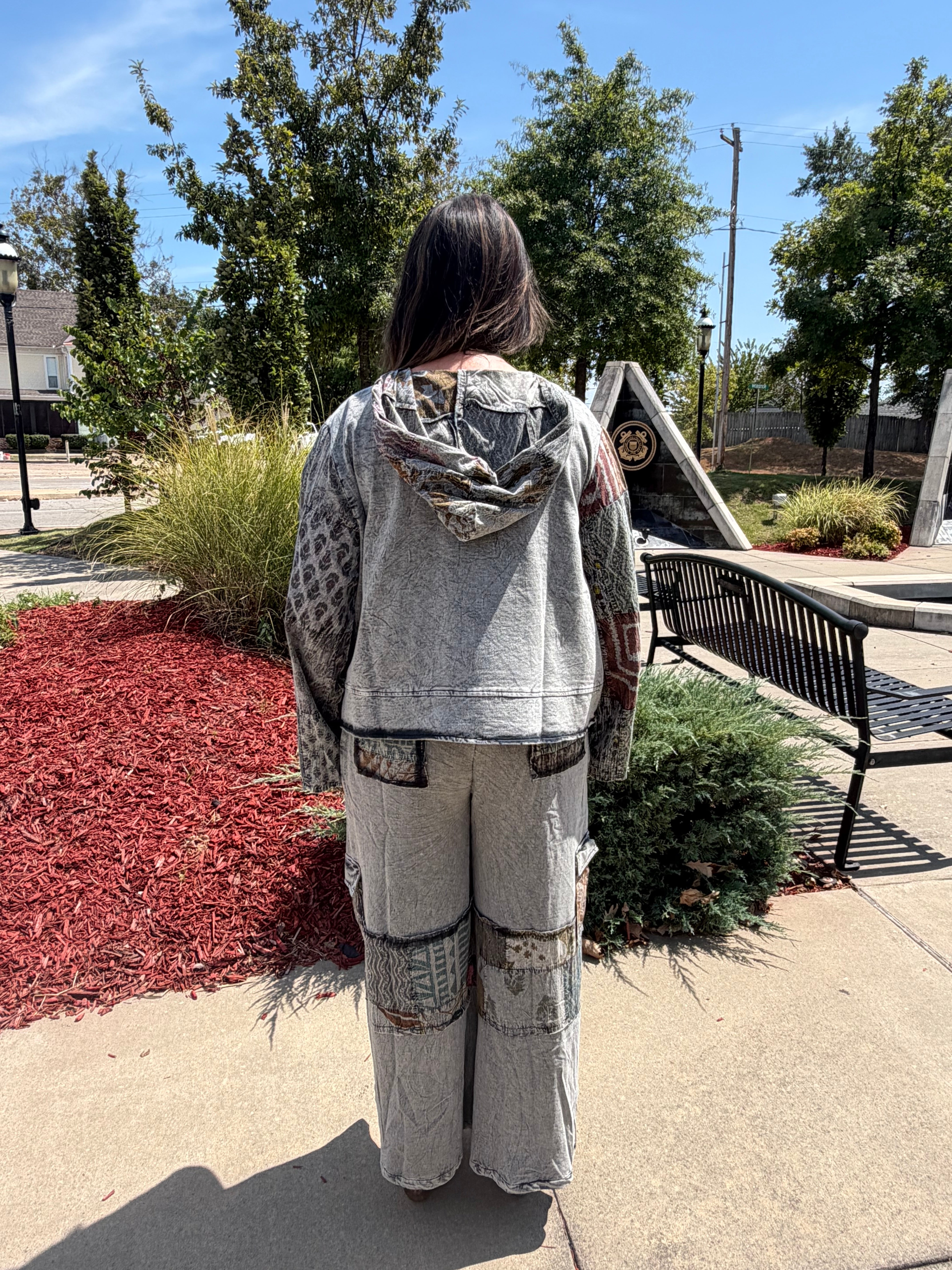 A person standing outdoors on a paved walkway, viewed from behind, wearing a light gray hooded sweatshirt and matching light gray patchwork wide-leg pants with darker gray and black distressed details.