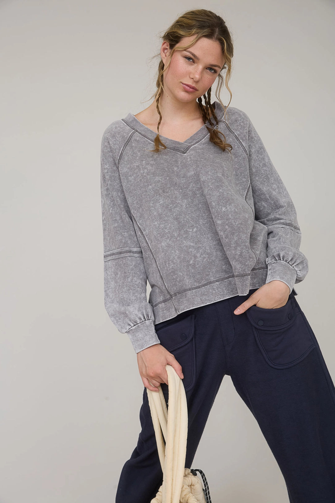Woman wearing a gray sweater and navy pants holding a beige bag against a plain background