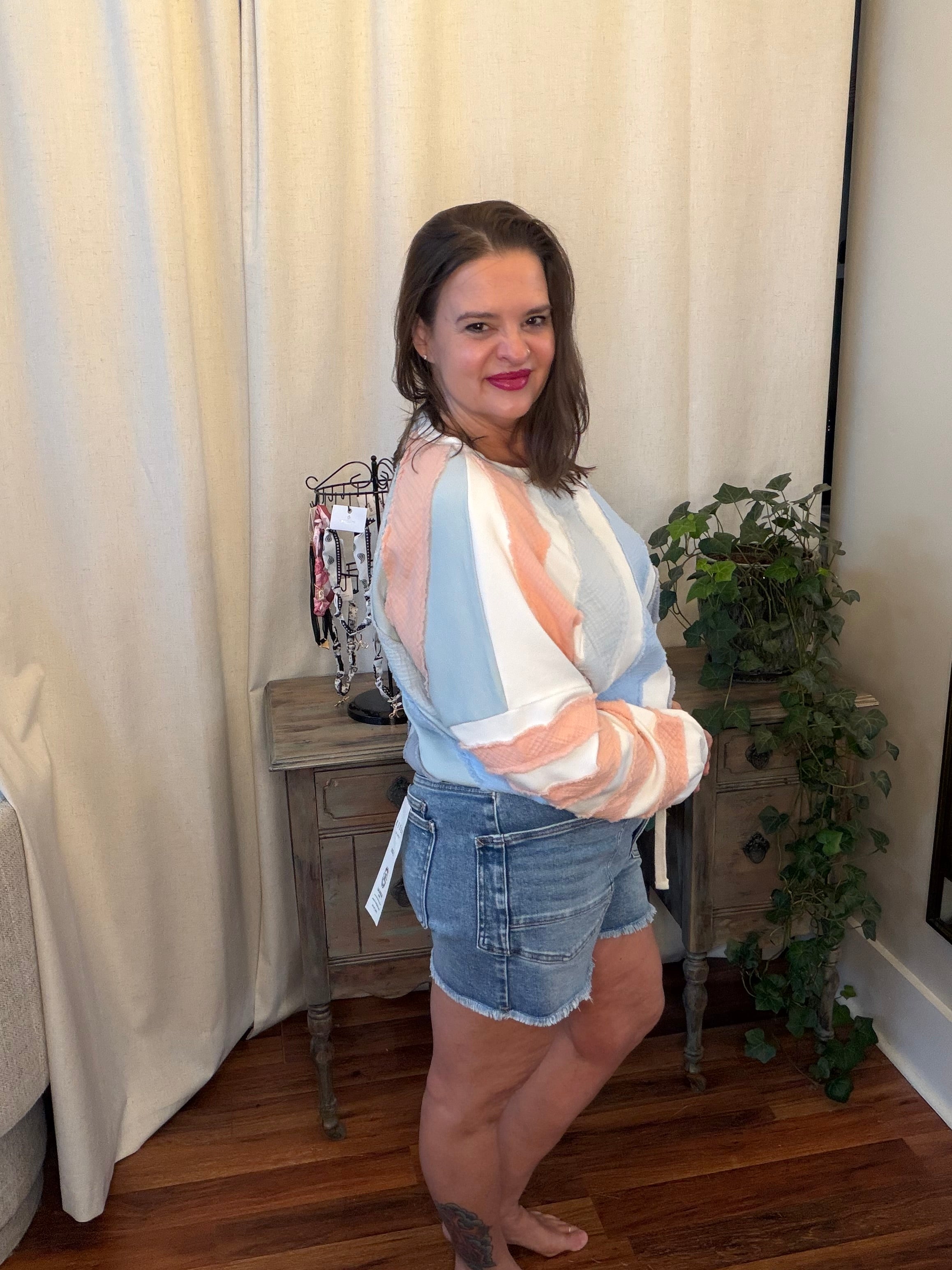 Woman wearing a striped shirt and denim shorts standing in a room with curtains and a plant.