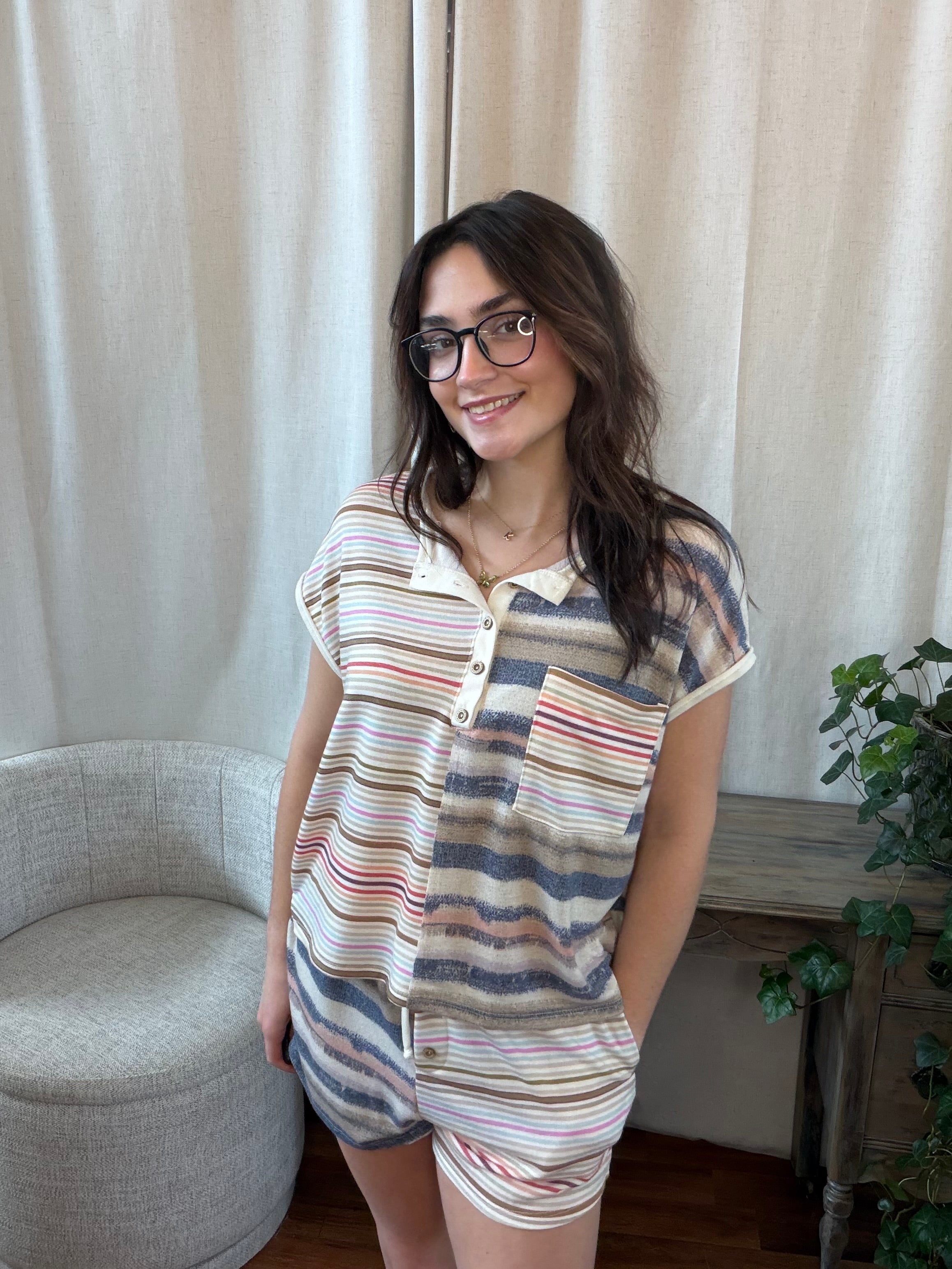 Woman wearing a striped shirt and shorts standing in a room with curtains and a plant.