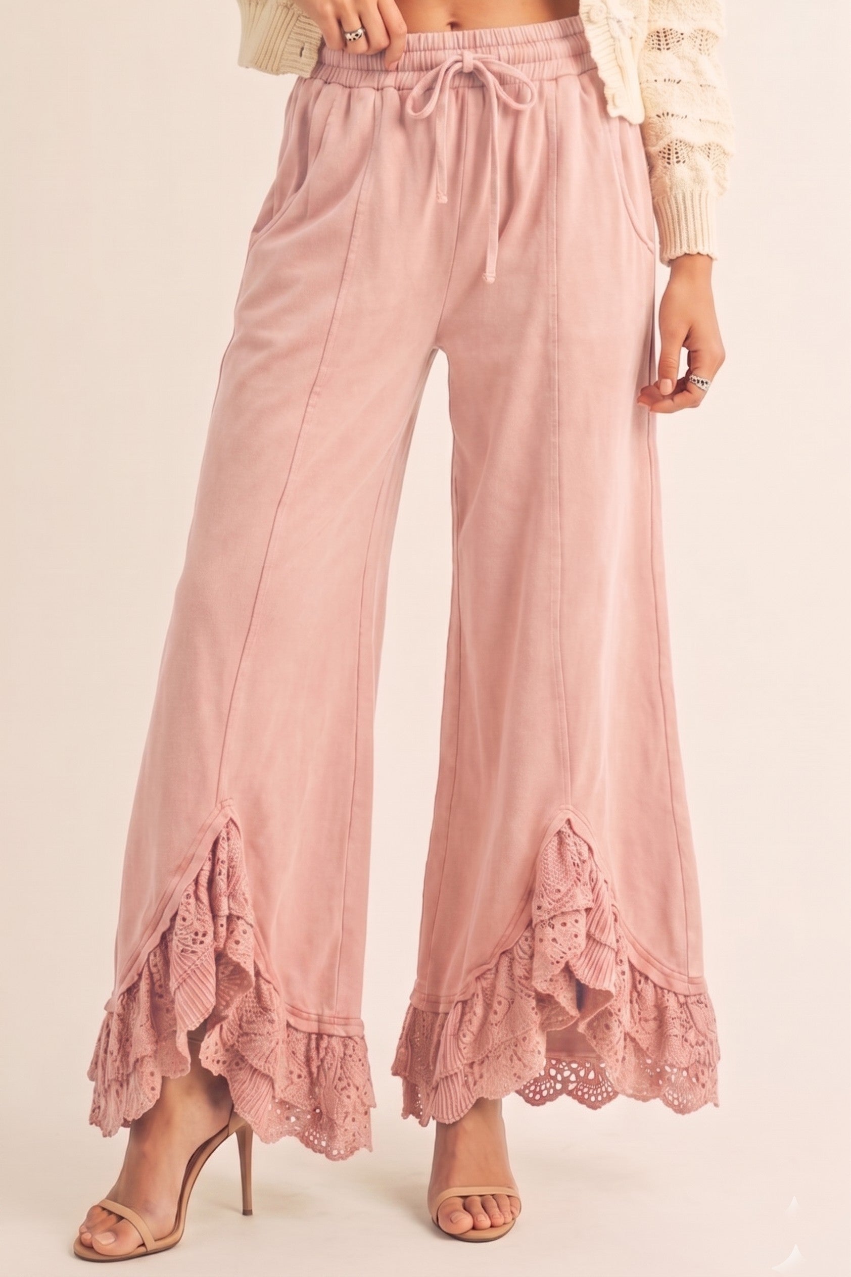 A person wearing dusty pink wide-leg drawstring pants with ruffled lace trim at the hem, paired with a cream knit top and nude high-heeled sandals, against a plain light beige studio background.