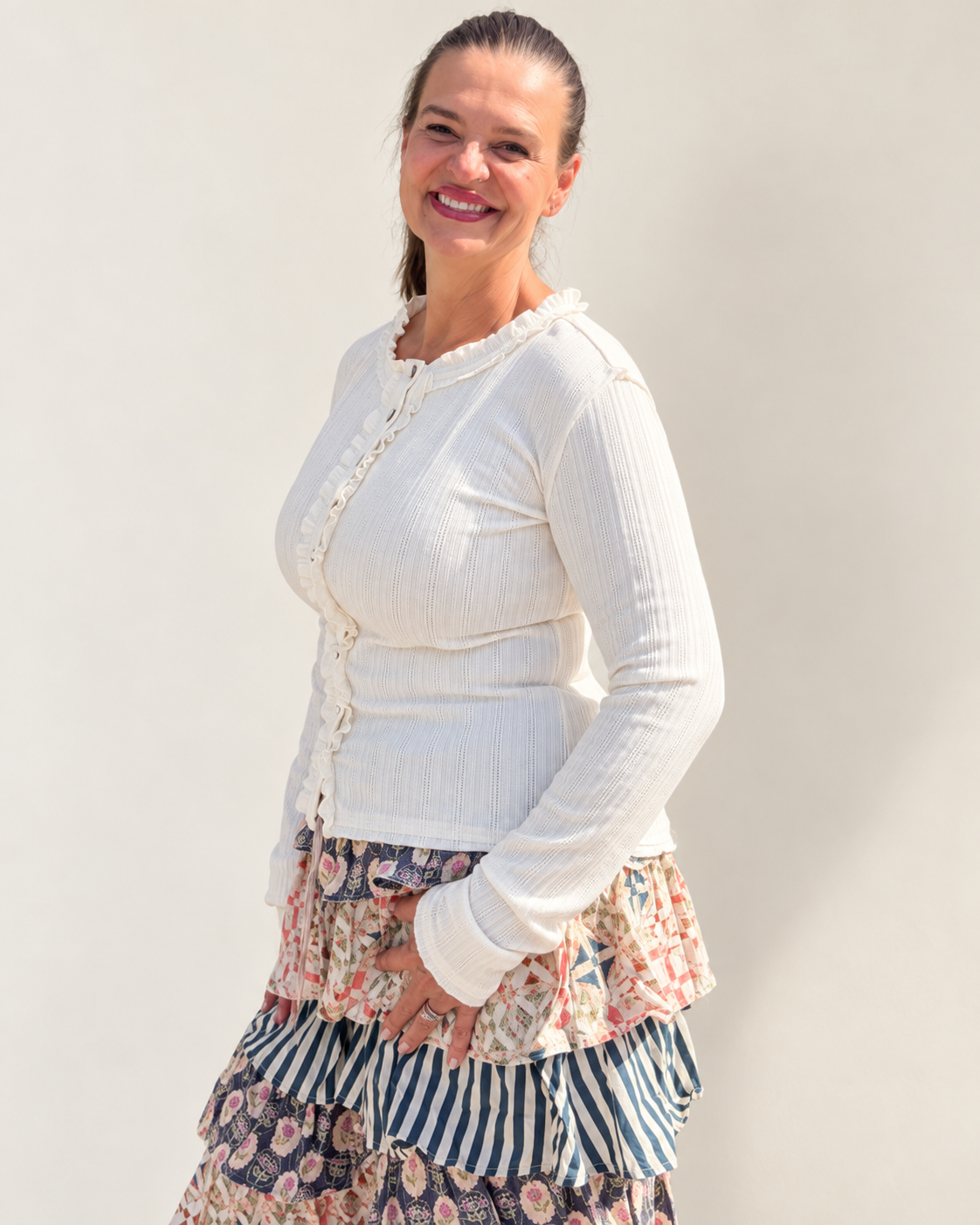 A woman modeling a white ribbed long-sleeve cardigan with ruffled trim over a tiered floral skirt, standing against a plain light beige wall.