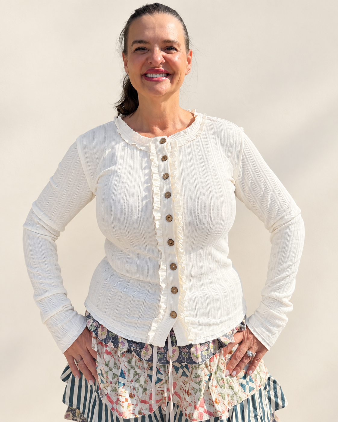 A woman wearing a cream-colored, long-sleeve crinkle-textured cardigan with brown buttons and a ruffled trim, paired with a colorful floral and striped skirt, posing against a plain beige background.