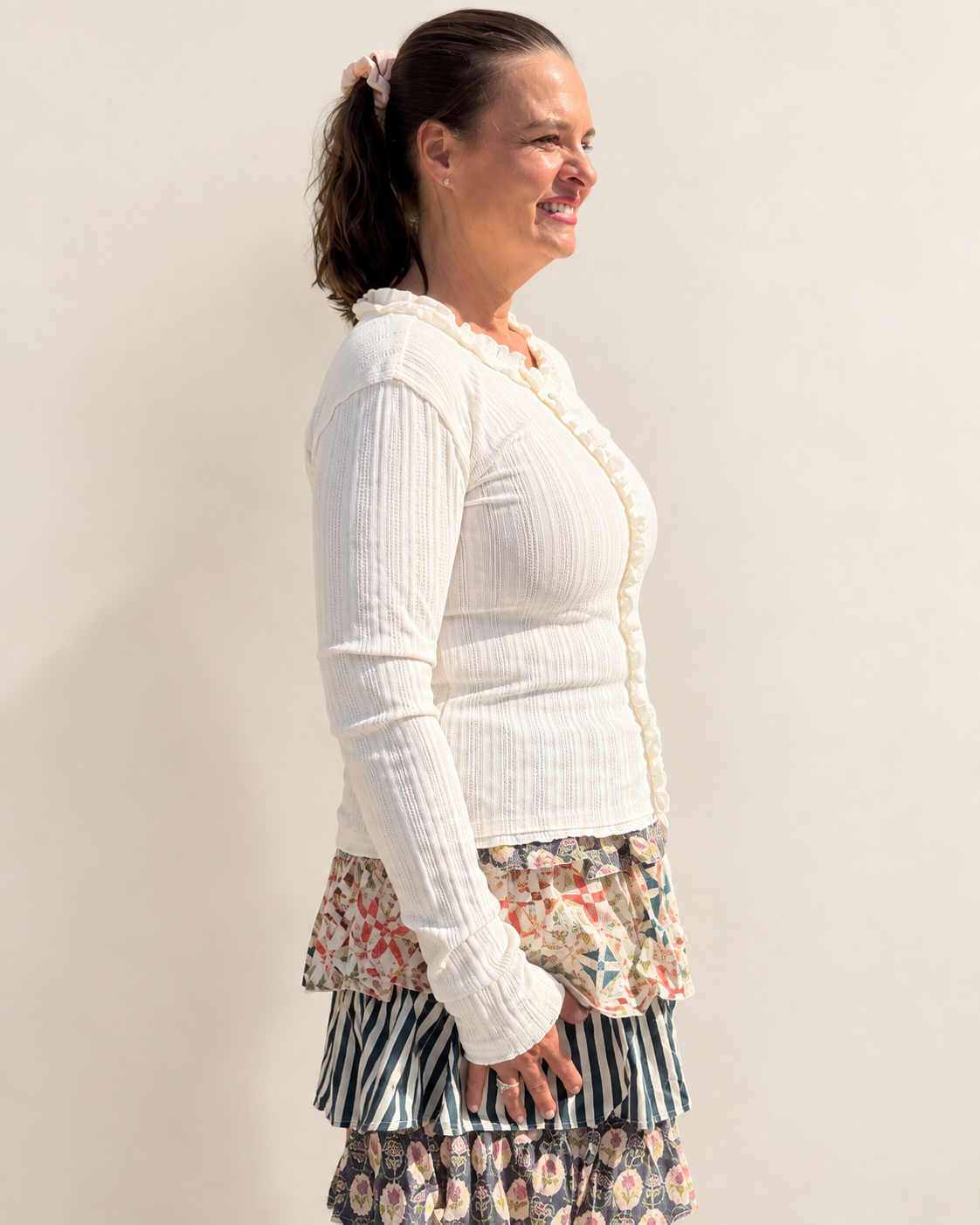 A woman shown in side profile wearing a white ribbed cardigan with a ruffled trim over a floral and striped layered skirt, against a plain off-white background.