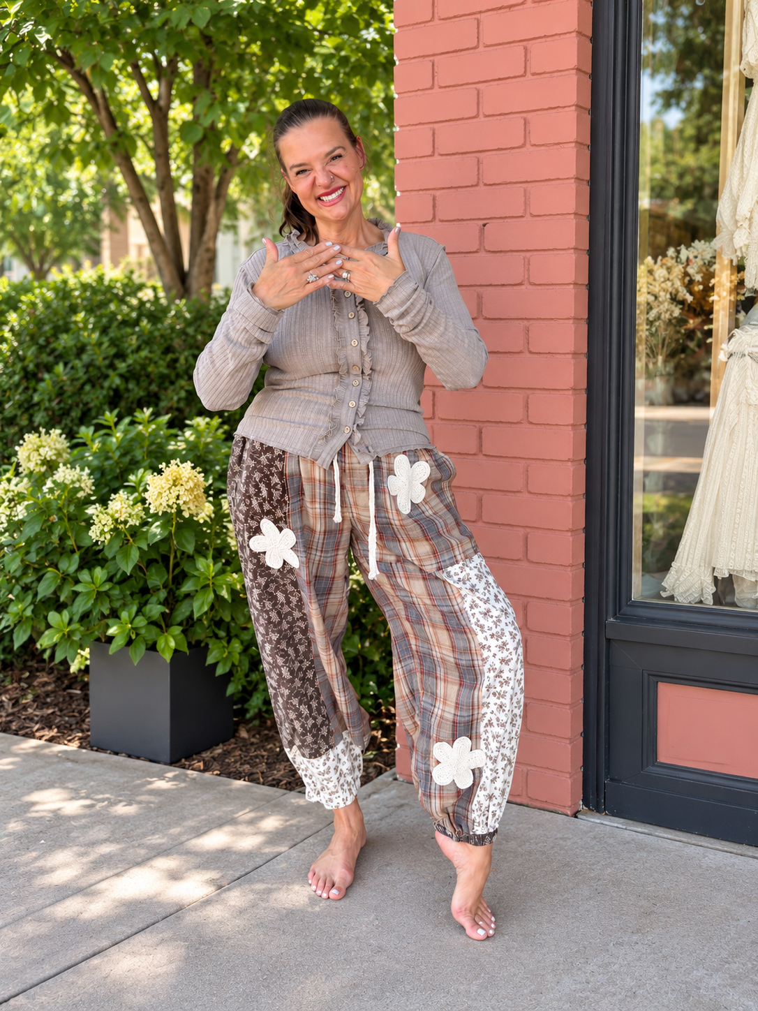 A woman stands barefoot outdoors on a sidewalk next to a brick building, wearing a gray button-front cardigan and patchwork pajama pants with floral appliques and plaid panels.