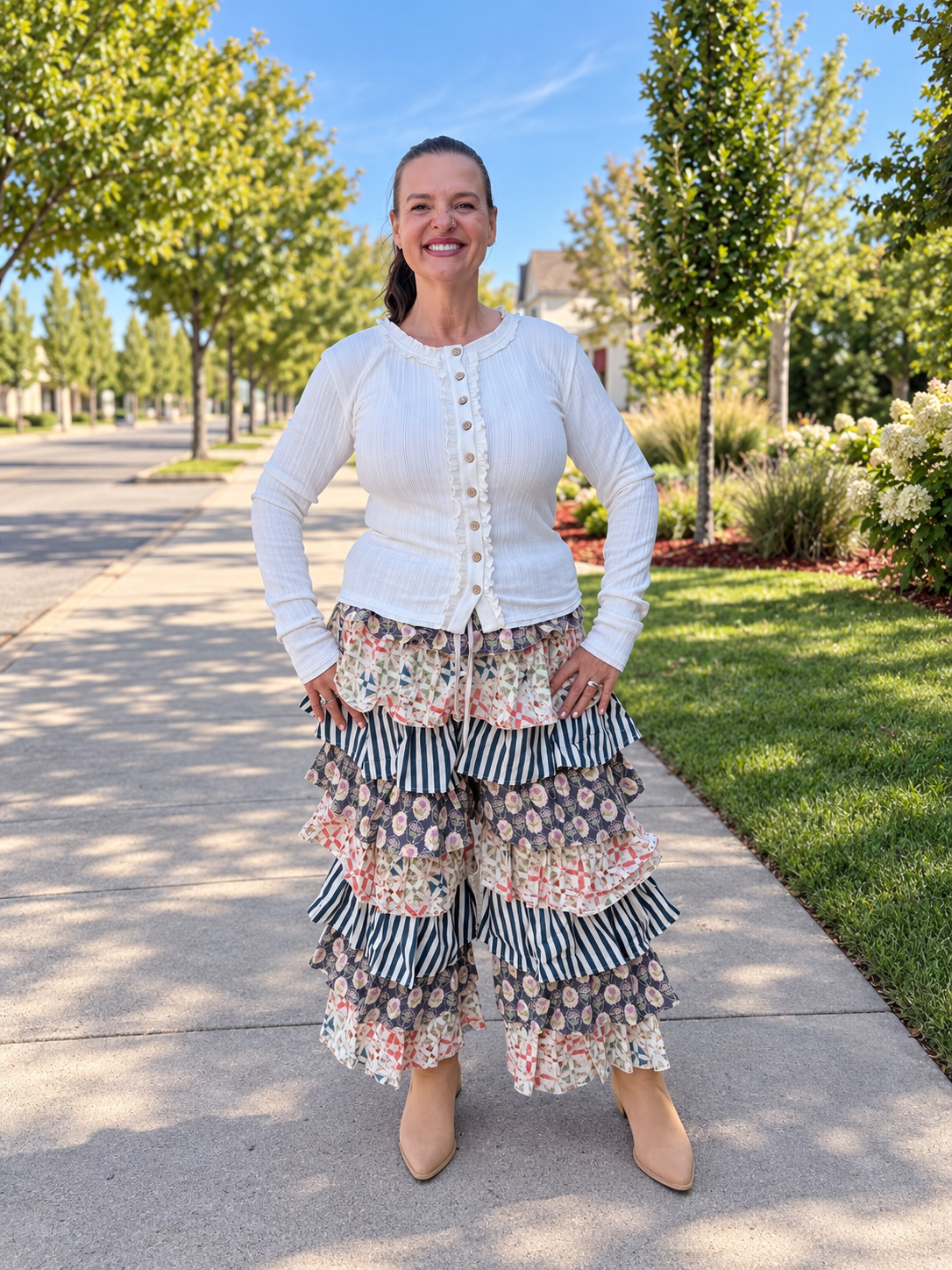 A woman stands on a paved outdoor walkway wearing a white long-sleeve button-front cardigan and a tiered ruffled pants from blue velvet with floral and striped patterns. Trees, grass, and a blue sky are visible in the background.