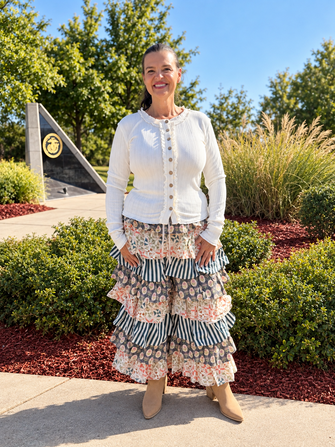 A woman stands outdoors in front of trees and landscaping, wearing a white long-sleeve top and a long tiered ruffled pants from bluevelvet with striped and floral patterns. 