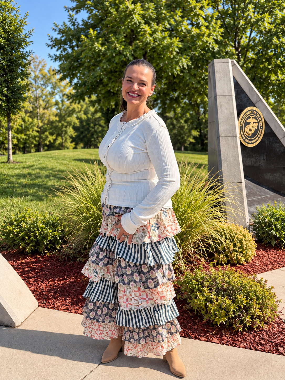 A woman stands outdoors in front of trees and landscaping, wearing a white long-sleeve top and a long tiered ruffled pants from bluevelvet with striped and floral patterns. 