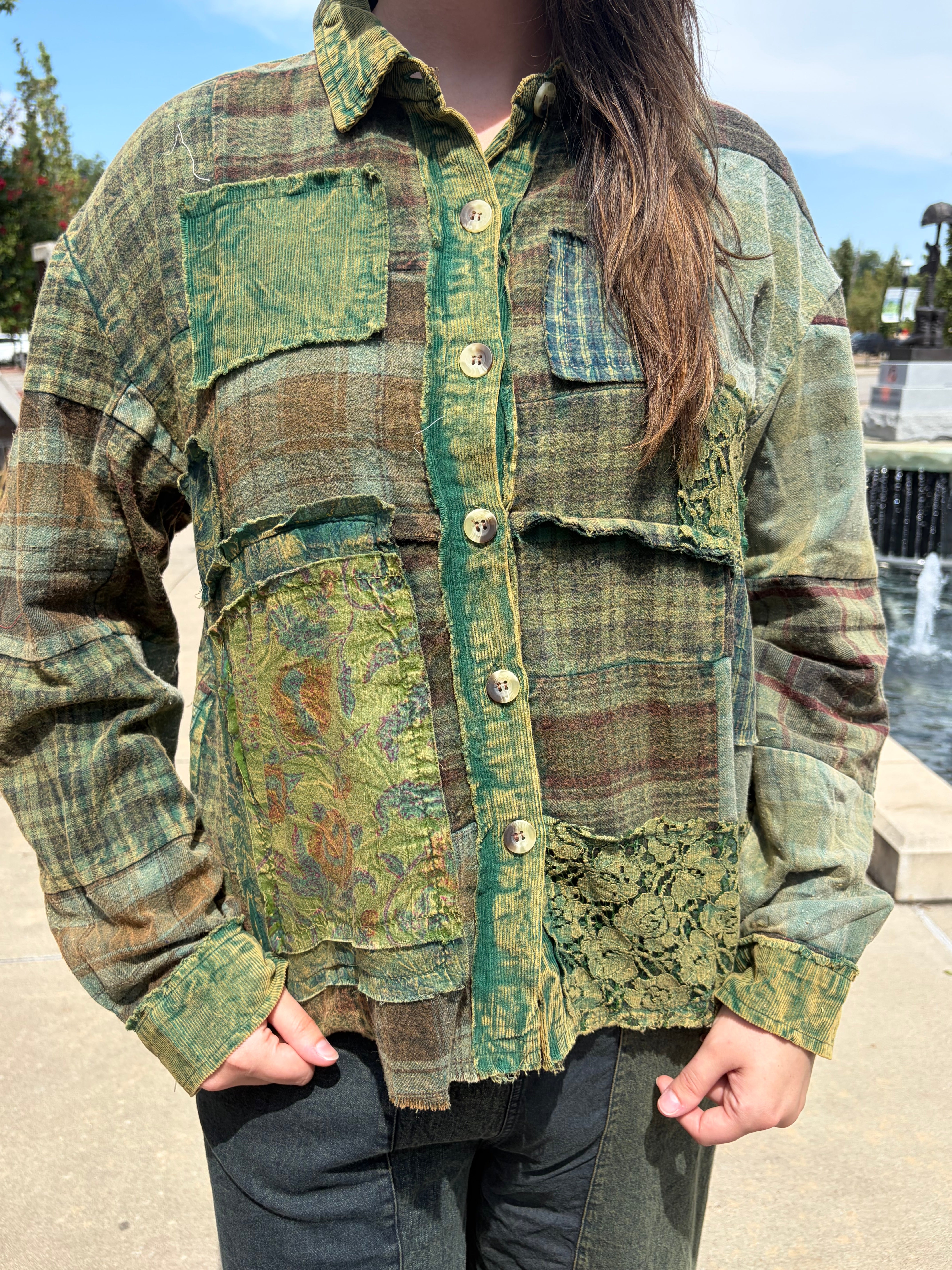 A person wearing a long-sleeve button-front patchwork shirt jacket in earthy green, brown, and tan tones, photographed outdoors near a fountain and trees.