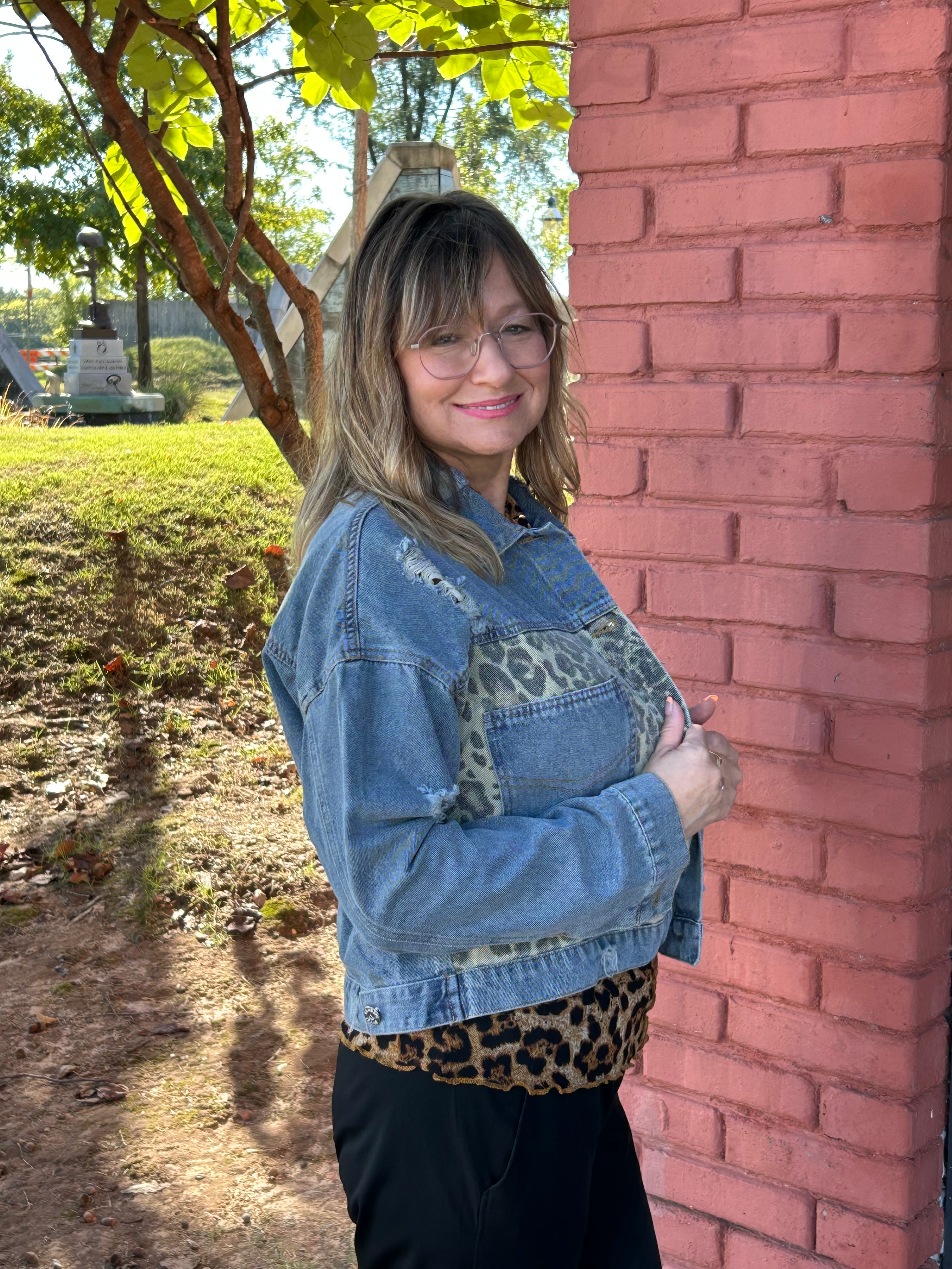 Woman wearing a denim jacket with leopard print accents against a brick wall.