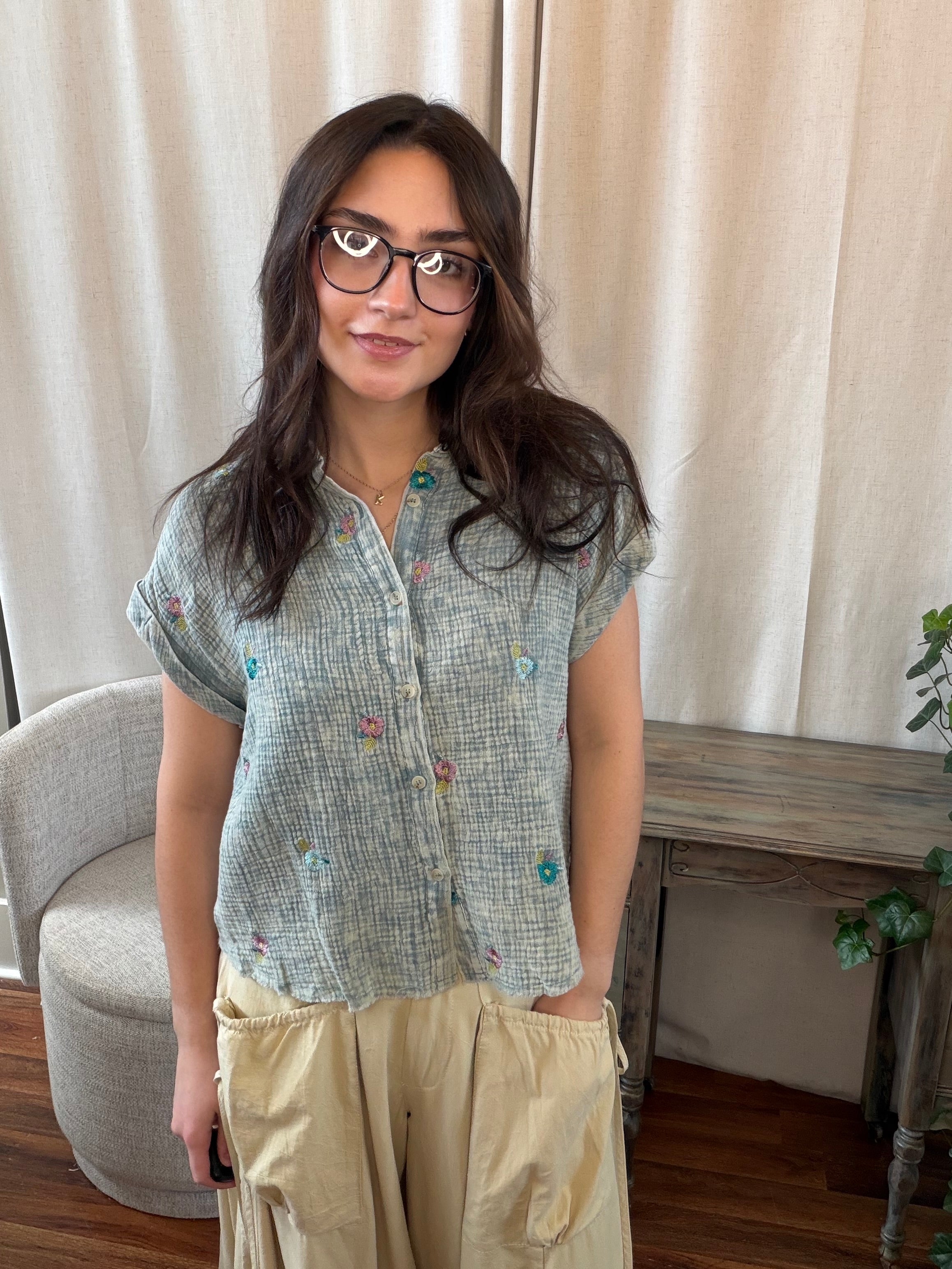 Woman wearing a patterned shirt and beige pants standing in a room with curtains and a table.