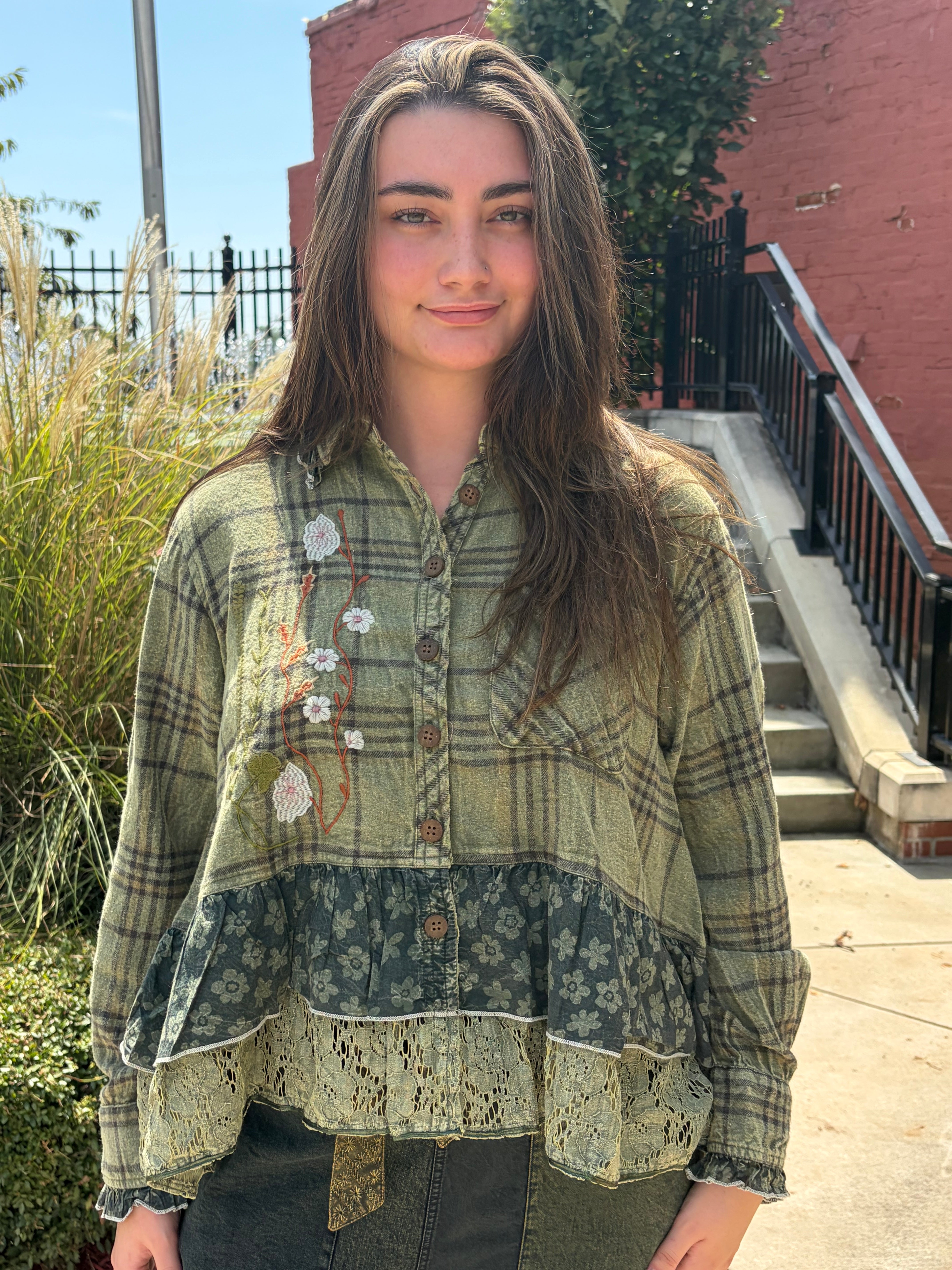 A woman wearing a long-sleeve patchwork button-up shirt with a green and brown plaid pattern, floral lace trim, and embroidered white flowers, standing outdoors in front of a brick building and staircase.