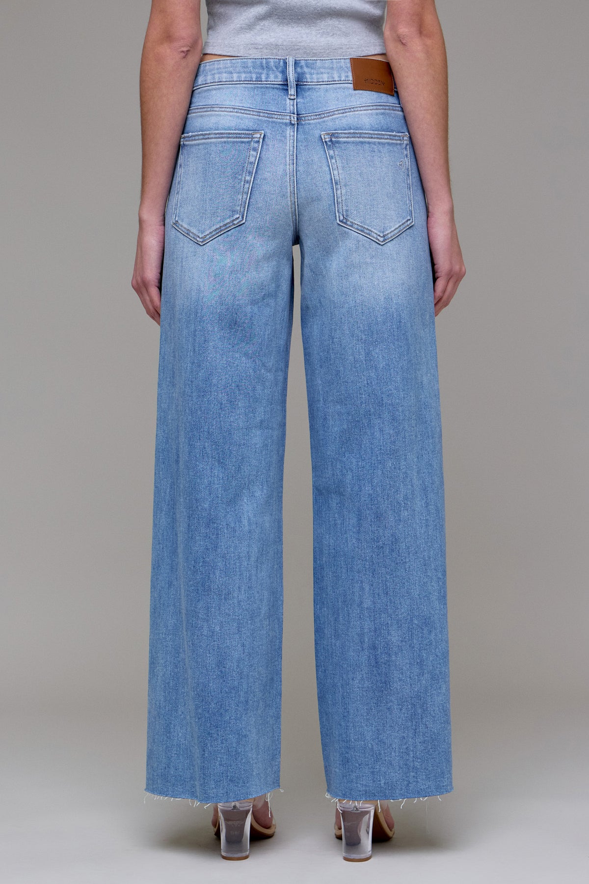 Back view of a model wearing Easygoing Edge medium-light wash wide-leg jeans with two back pockets, a brown waistband patch, and raw hems.
