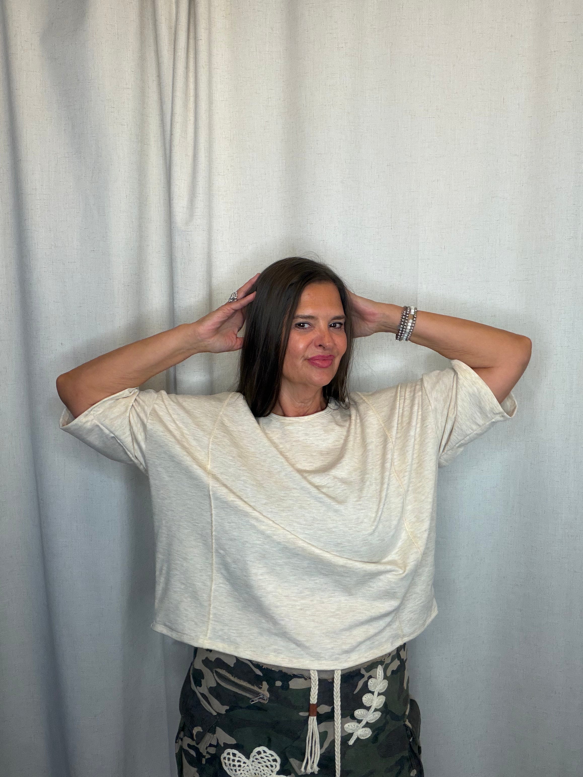 A woman posing in front of a light gray curtain, wearing a cream-colored draped short-sleeve t-shirt and camouflage drawstring pants.