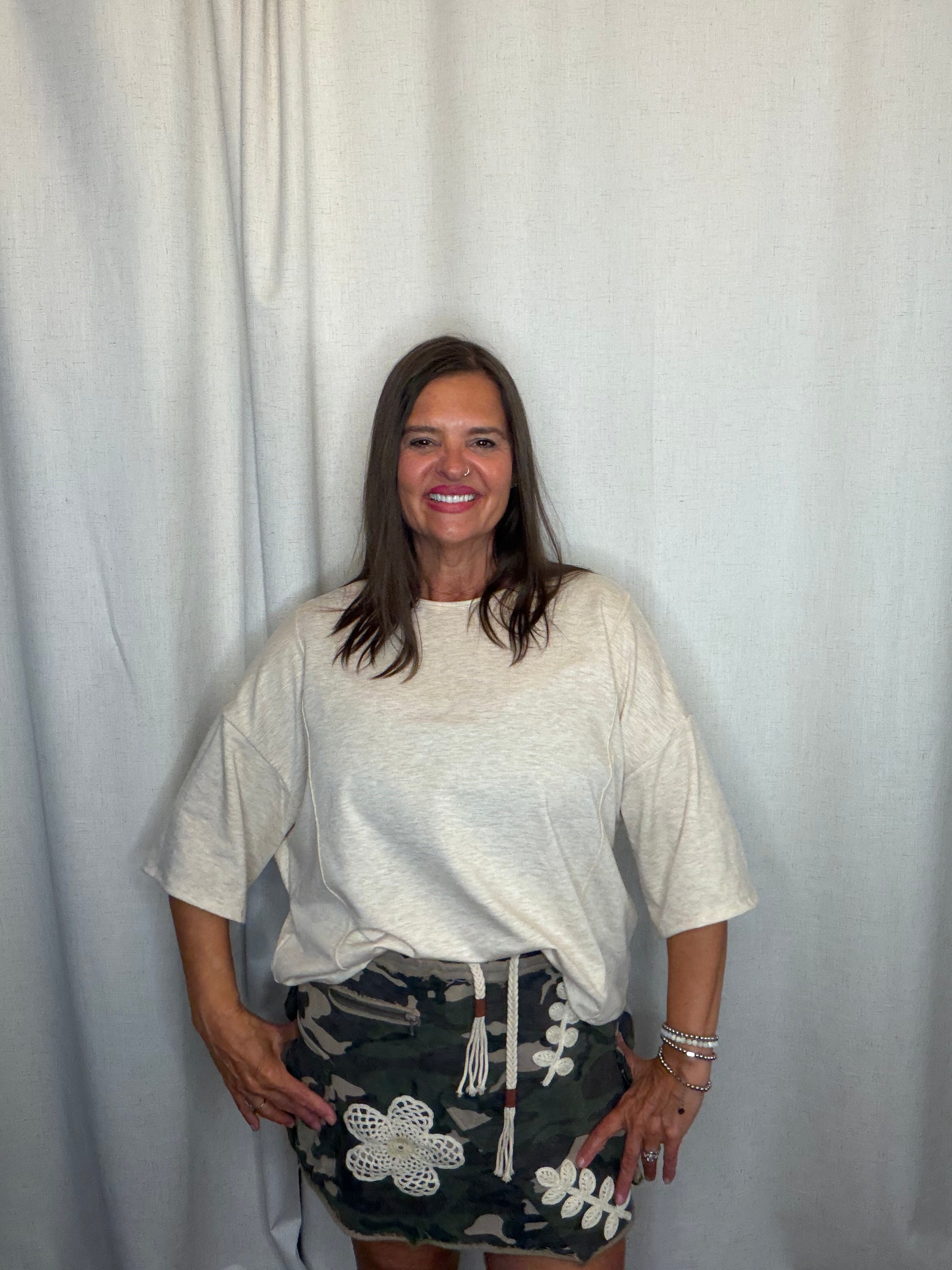 A woman posing in front of a white curtain backdrop, wearing an oversized off-white t-shirt and a dark green camouflage mini skirt with white crochet flower details.