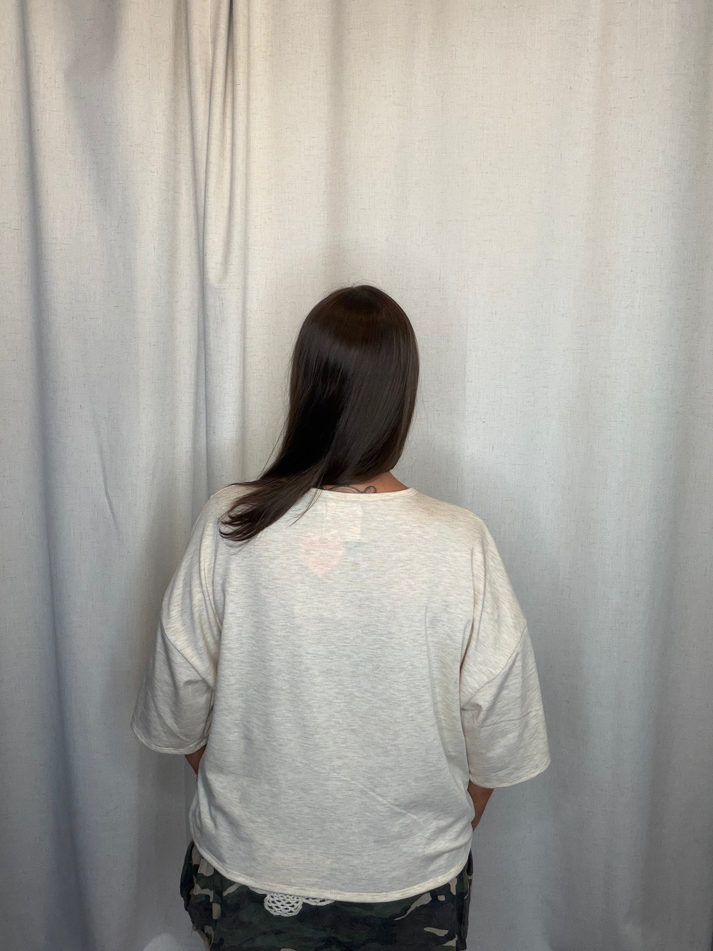 A person viewed from behind wearing a light heather gray oversized short-sleeve t-shirt and dark camouflage pants, standing in front of a light gray fabric backdrop.