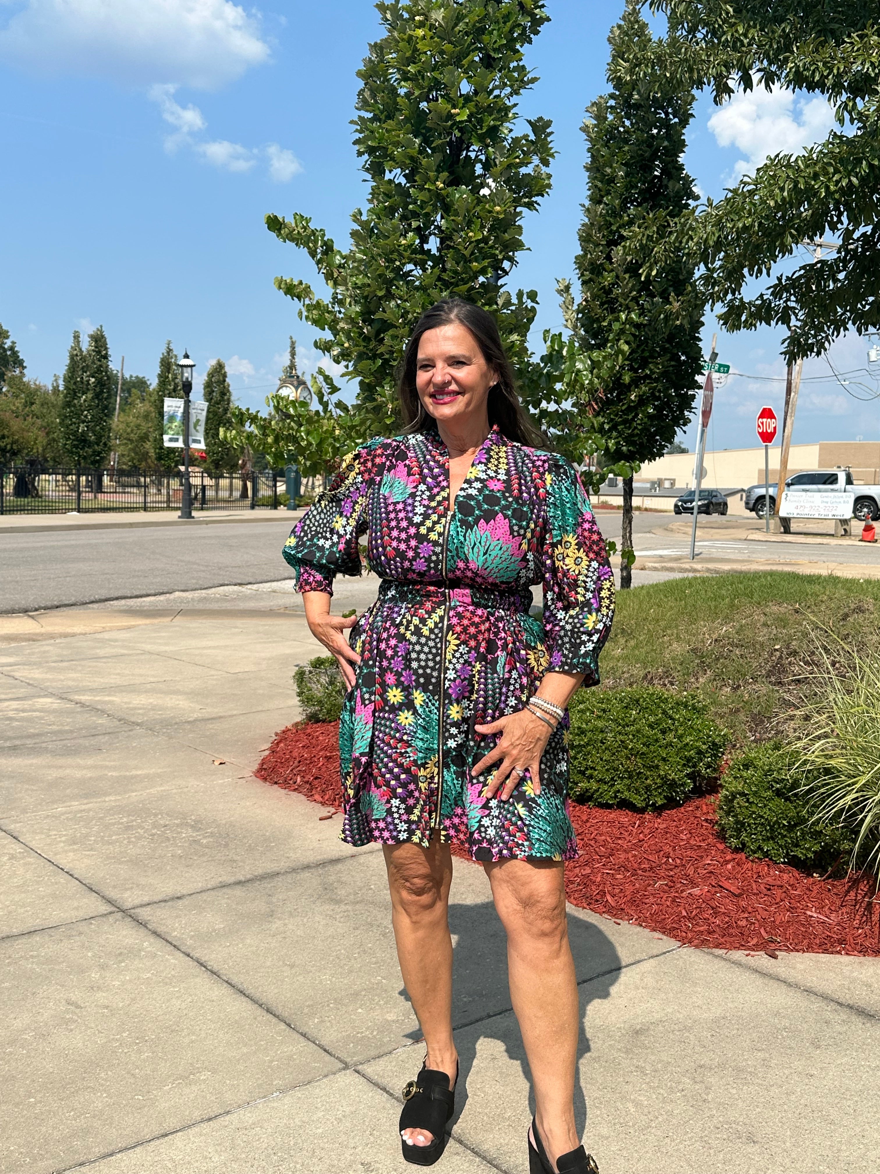 A woman stands outdoors on a sidewalk wearing a short puff-sleeve dress with a colorful geometric pattern in purple, green, pink, yellow, and blue. She is wearing black platform sandals, with trees, a street, and a parking lot visible in the background.