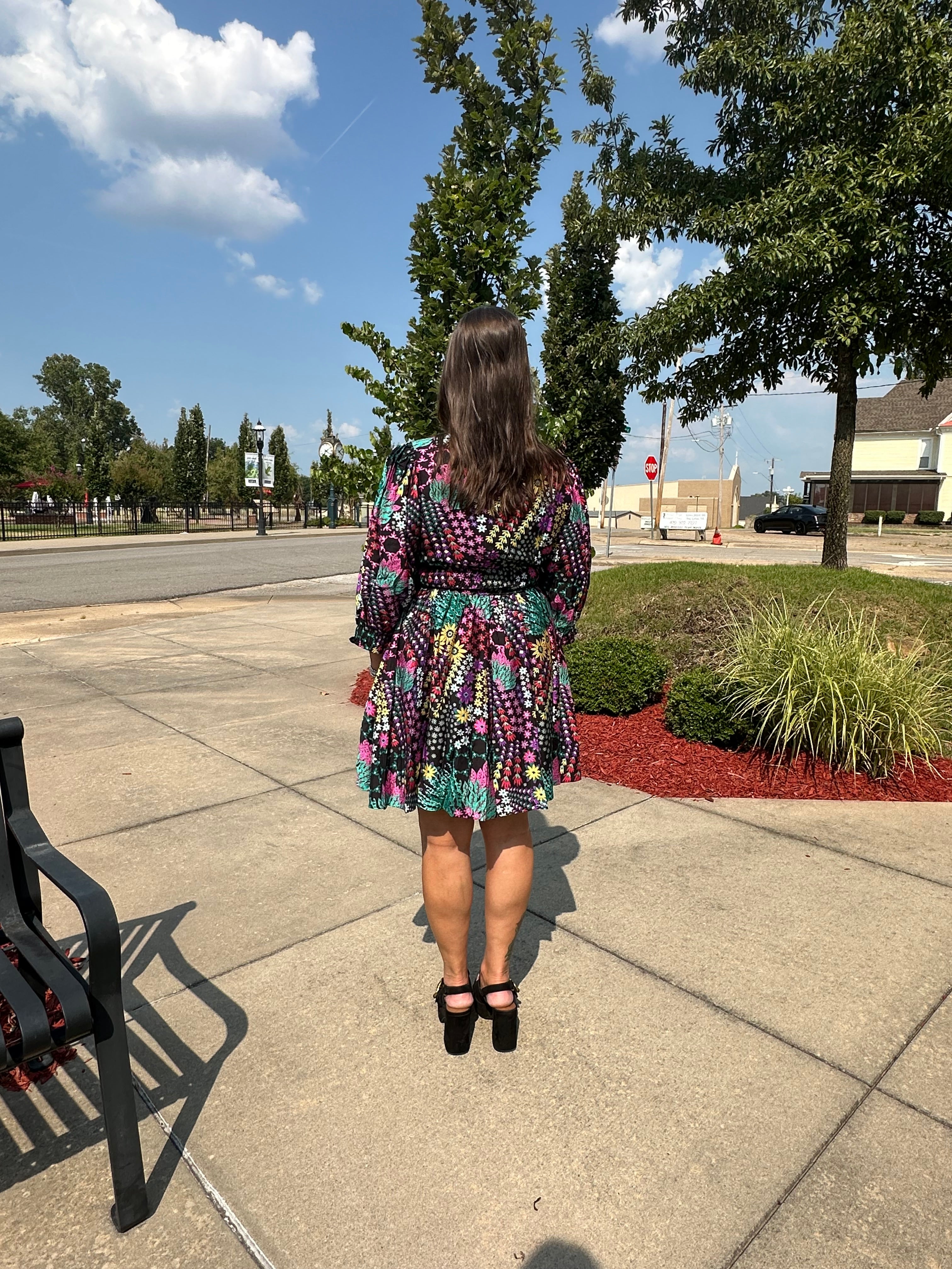 A woman viewed from behind wearing a colorful, long-sleeved mini dress with a black and white polka dot pattern and black high heels, standing on a concrete sidewalk outdoors.