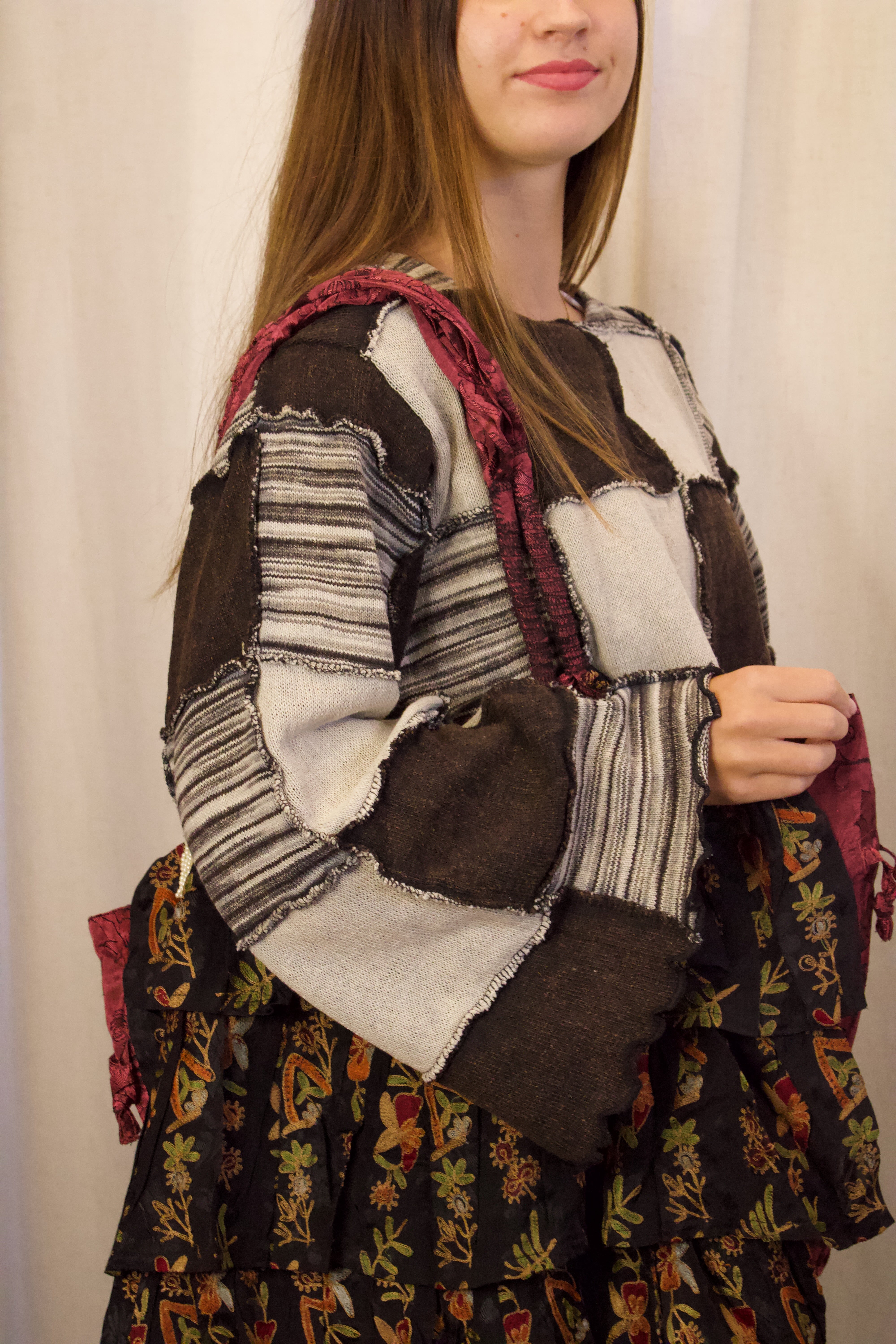 A woman wearing a bohemian patchwork sweater and a dark floral tiered skirt, carrying a burgundy shoulder bag. The sweater features patchwork panels of brown, gray, and white striped wool.