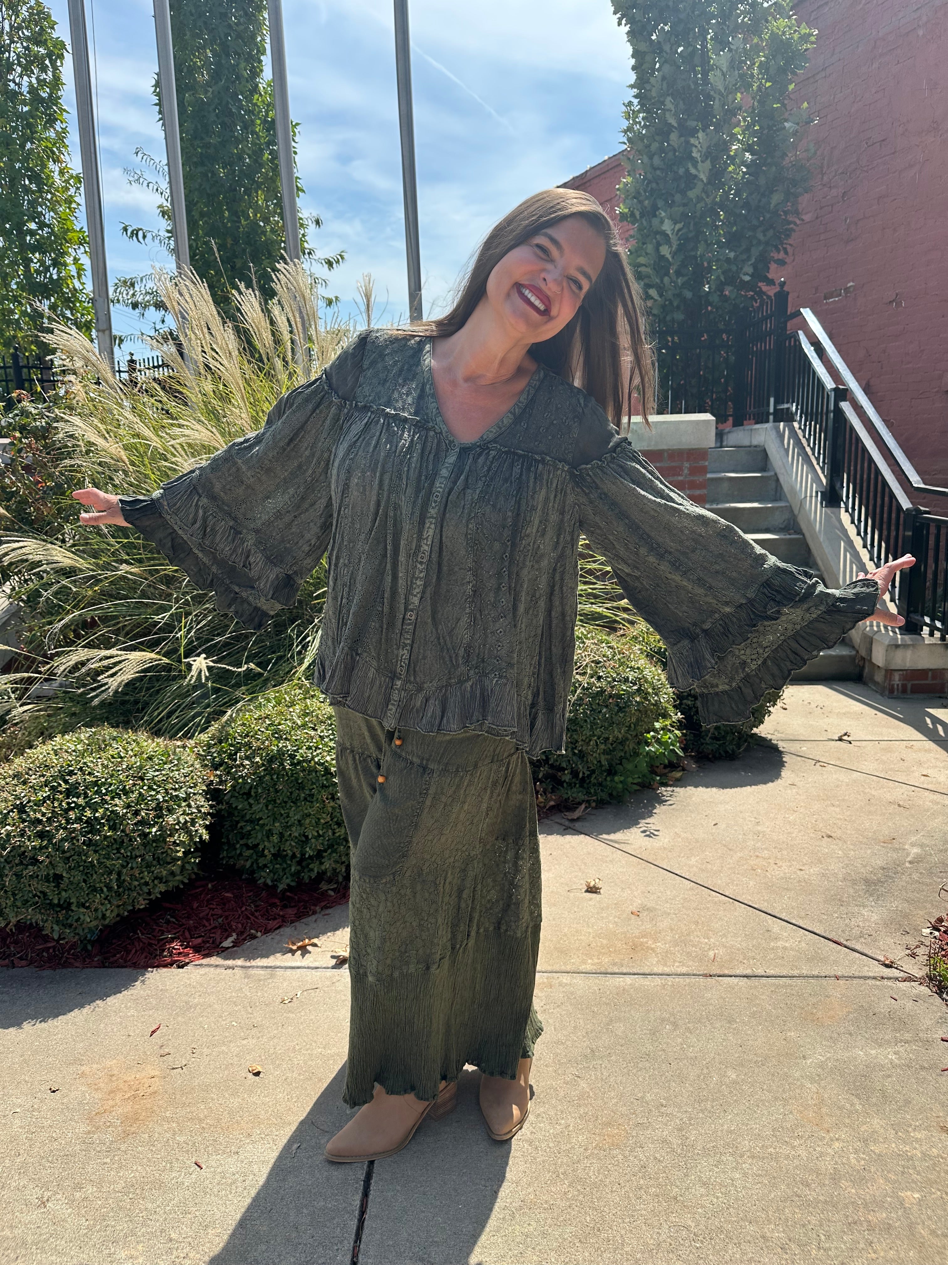 A woman wearing a matching olive green long-sleeve button-up shirt and wide-leg skirt stands outdoors on a concrete walkway with her arms outstretched, smiling broadly.