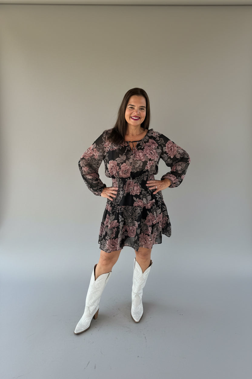 A woman posing, wearing a black long-sleeve mini dress with a dusty pink floral pattern and white cowboy boots.