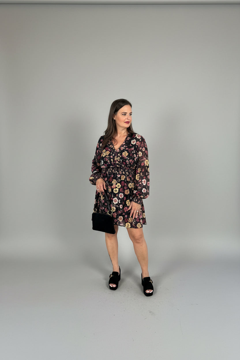 A woman stands under a covered outdoor walkway wearing a short floral print dress with long bell sleeves, black platform sandals, and holding a black clutch bag.