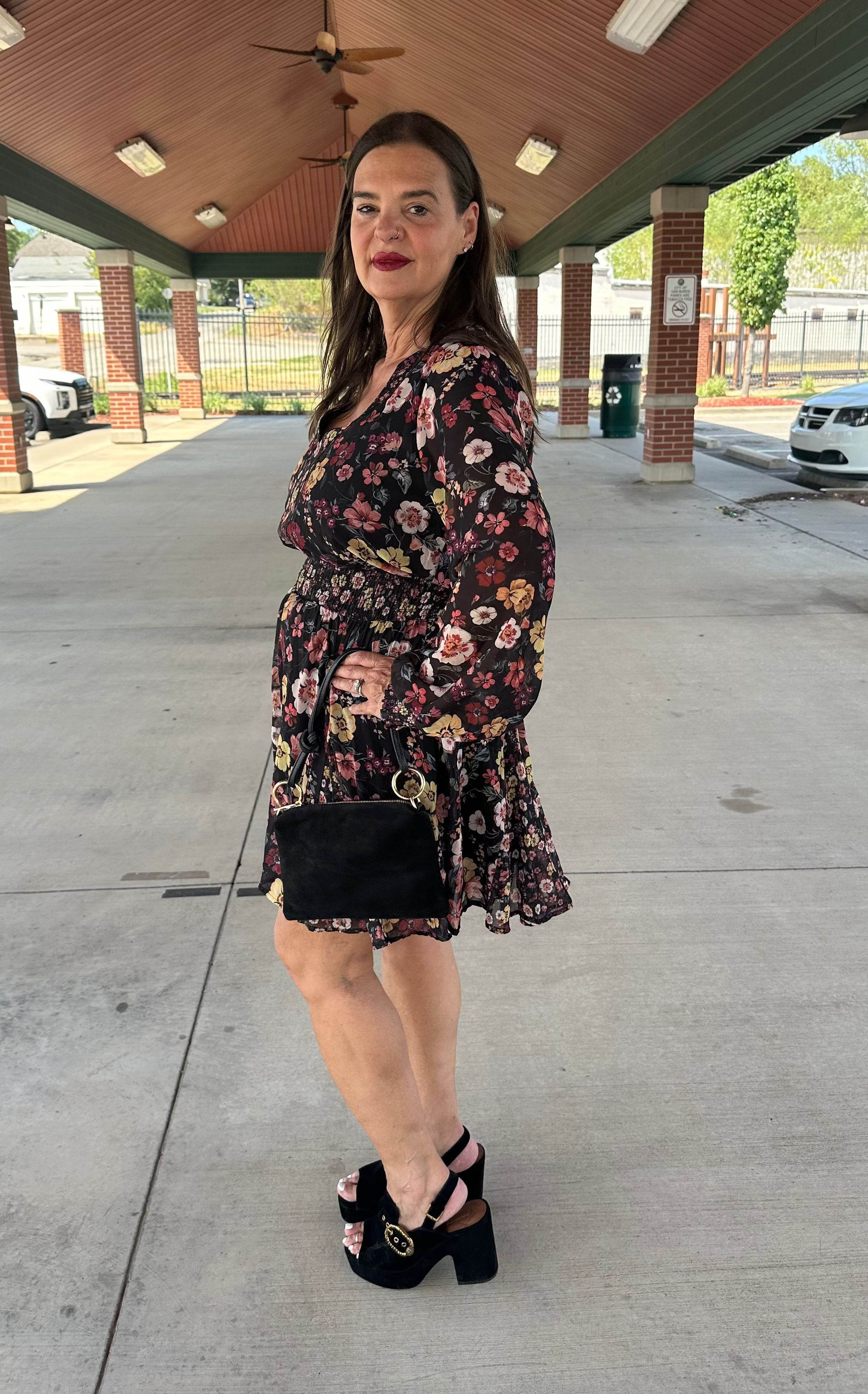 A woman stands under a covered outdoor walkway wearing a long-sleeve floral mini dress with a black base and multicolor print, holding a small black handbag and wearing black platform heels.