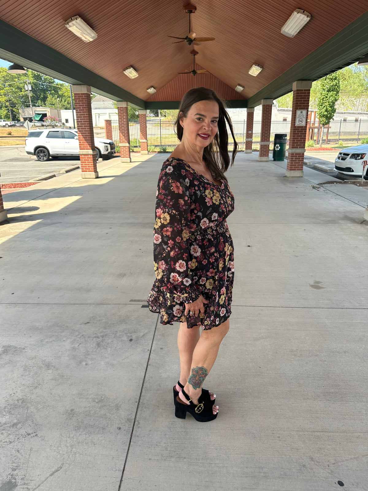 A woman wearing a black floral long-sleeve mini dress and black platform heels stands under a covered outdoor walkway with brick pillars and concrete flooring.