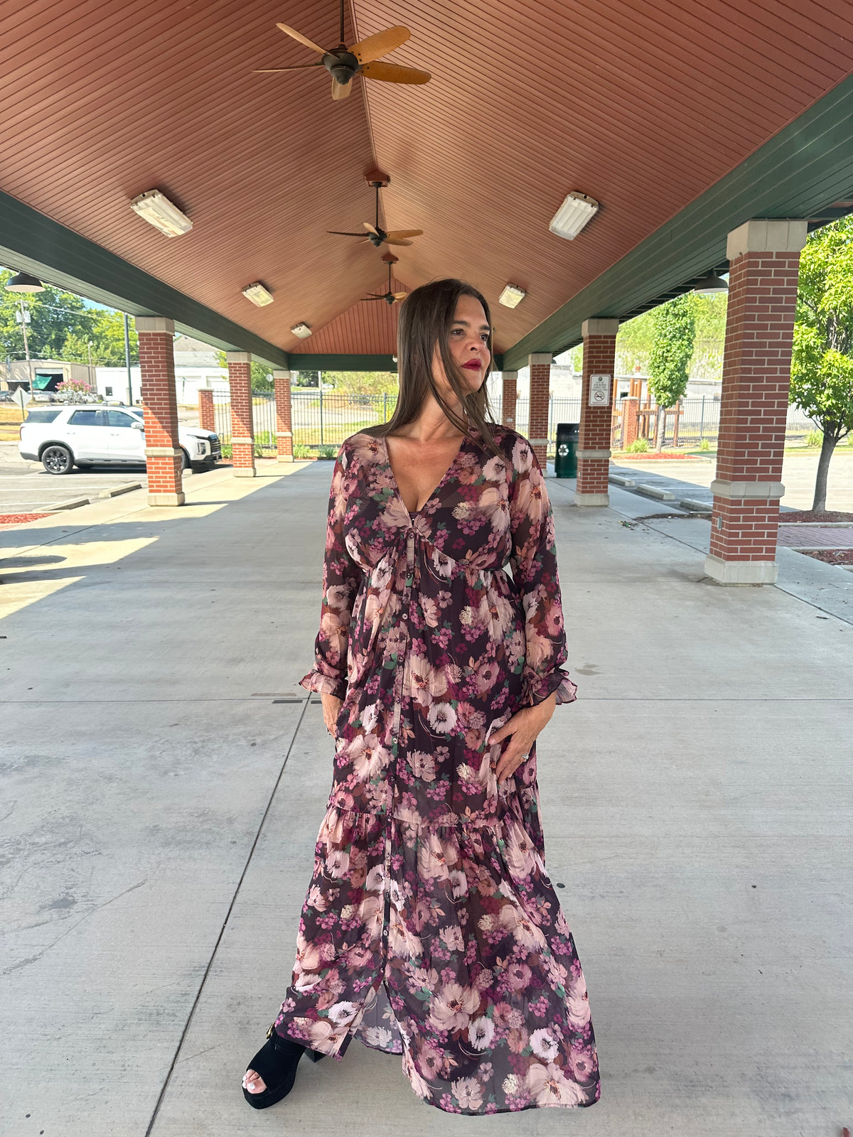 Woman wearing burgundy floral long-sleeve maxi dress with V-neckline and tiered hem, styled with black heeled sandals, outdoor pavilion setting
