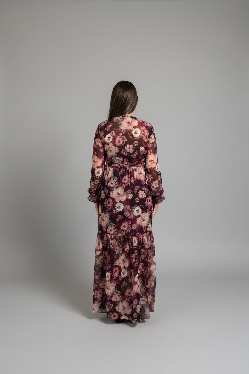 A woman viewed from behind wearing a long-sleeved, floor-length floral robe with a pink, purple, and black floral pattern, walking under a covered outdoor walkway.