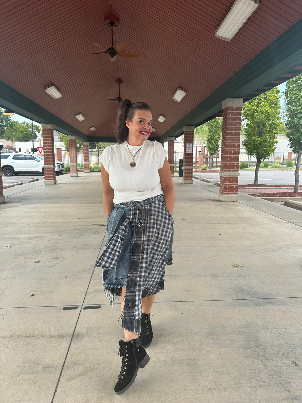 A woman stands under a covered walkway wearing a white short-sleeve t-shirt, a blue and gray plaid skirt tied around her waist, and black lace-up combat boots.