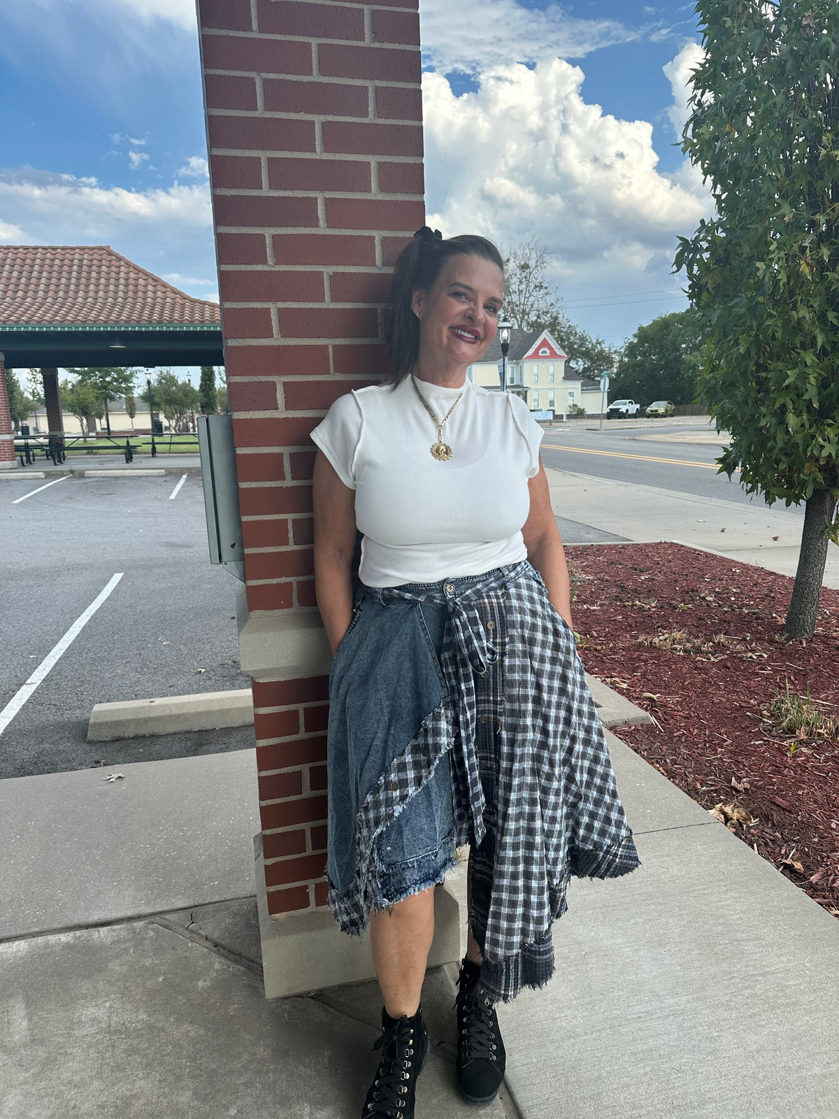 A woman stands outdoors against a red brick pillar wearing a white ruffled short-sleeve top, a black and white gingham shirt tied around her waist, a denim skirt, and black lace-up boots.