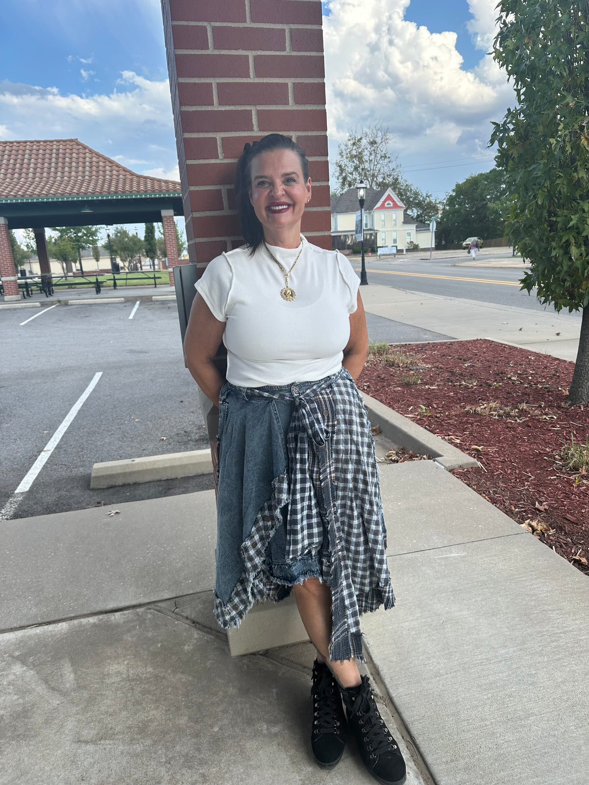 A woman posing outdoors against a brick pillar, wearing a white t-shirt, a blue denim skirt, and a black and white gingham shirt tied around her waist, paired with black lace-up boots.