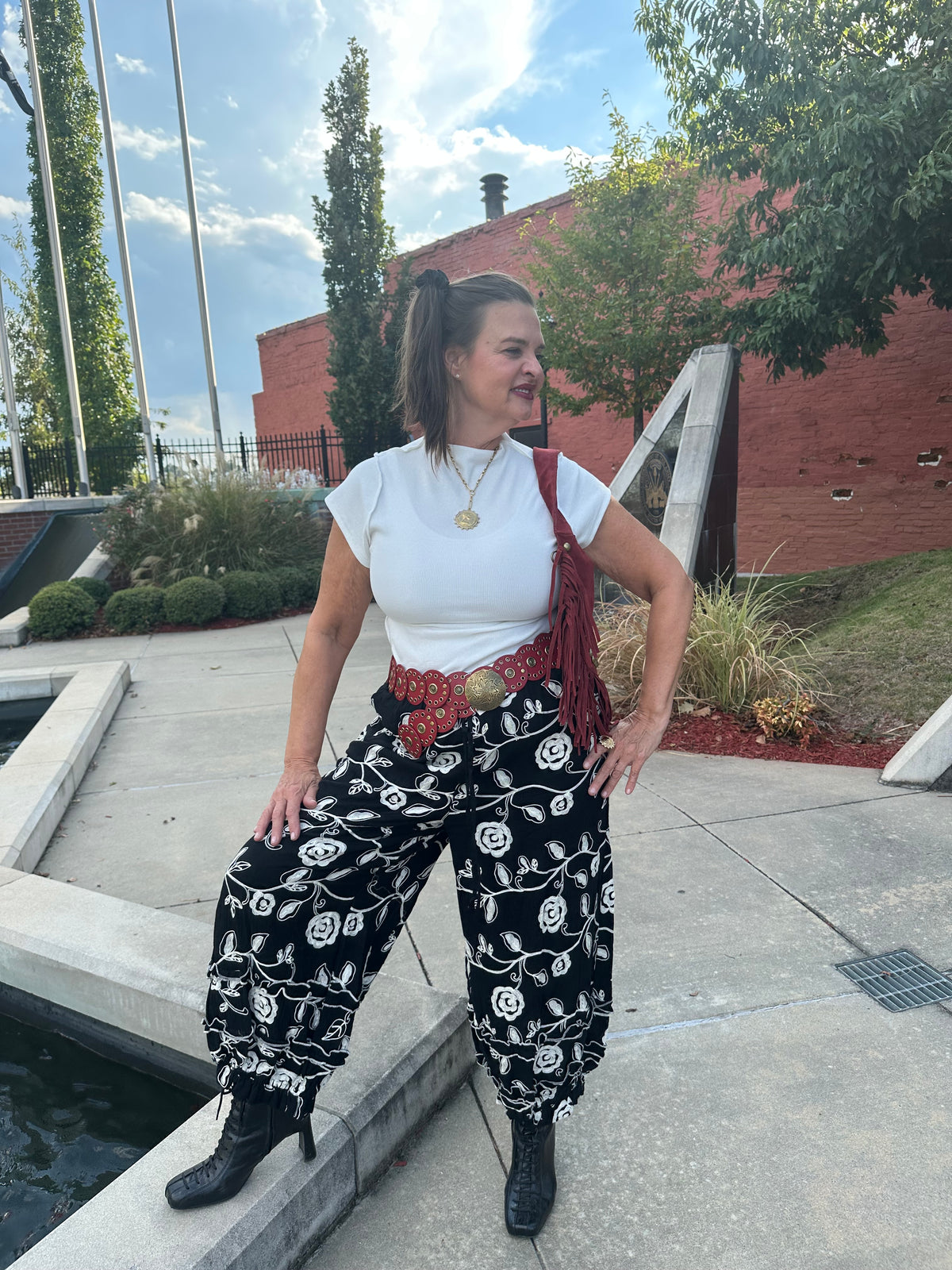 A woman wearing a white t-shirt, black and white patterned harem pants, a red fringed belt, and black boots, posing outdoors on a concrete plaza next to a water feature.