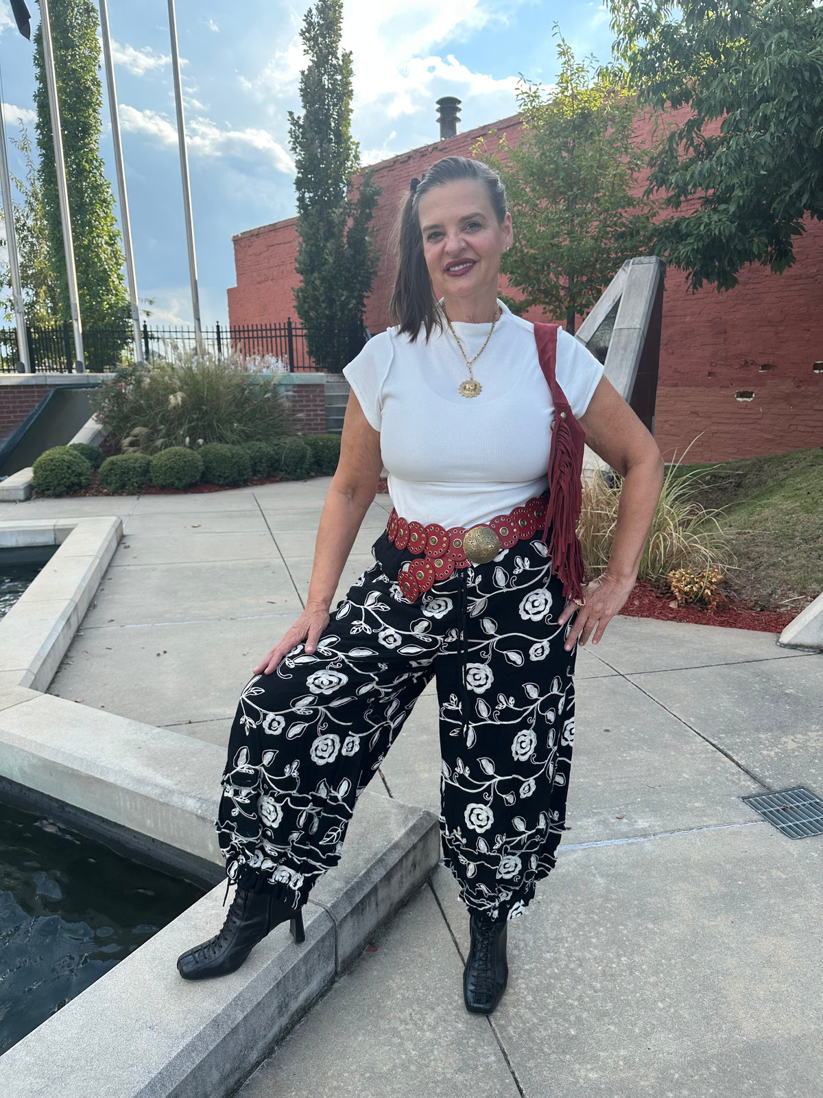 A woman posing outdoors in a white t-shirt, black harem-style pants with a white rose pattern, a red patterned belt, and black boots, standing next to a concrete pool.