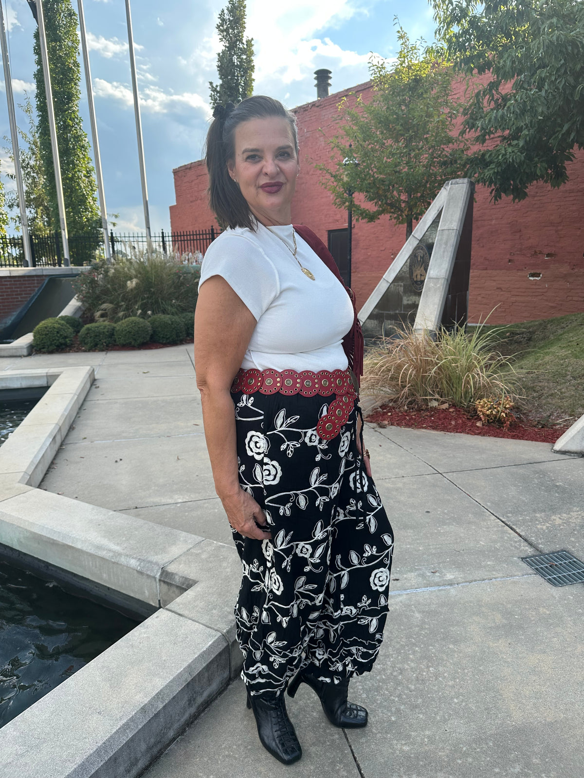 A woman wearing a white short-sleeve top, a red patterned belt, and black wide-leg pants with a white floral print, standing outdoors on a concrete plaza next to a water feature.