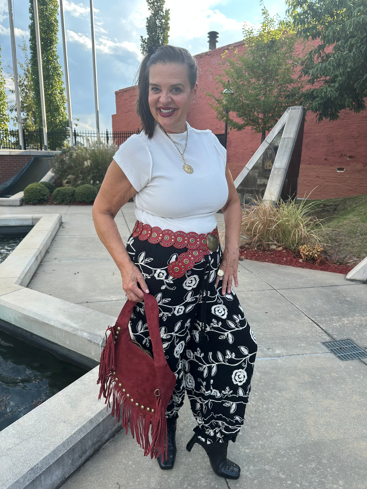 A woman stands outdoors wearing a white short-sleeve top, black wide-leg pants with a white floral vine print, a red patterned belt, black ankle boots, and holding a red fringe handbag.