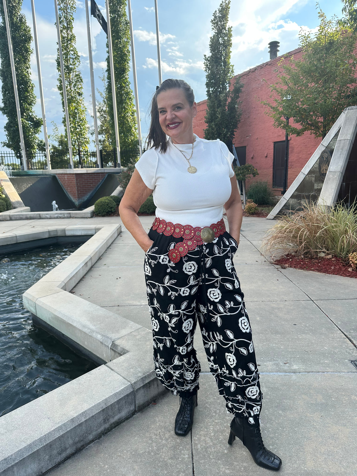 A woman stands outdoors on a concrete walkway next to a water feature, wearing a white short-sleeve top, black wide-leg pants with a white floral vine pattern, a red patterned belt, and black ankle boots.