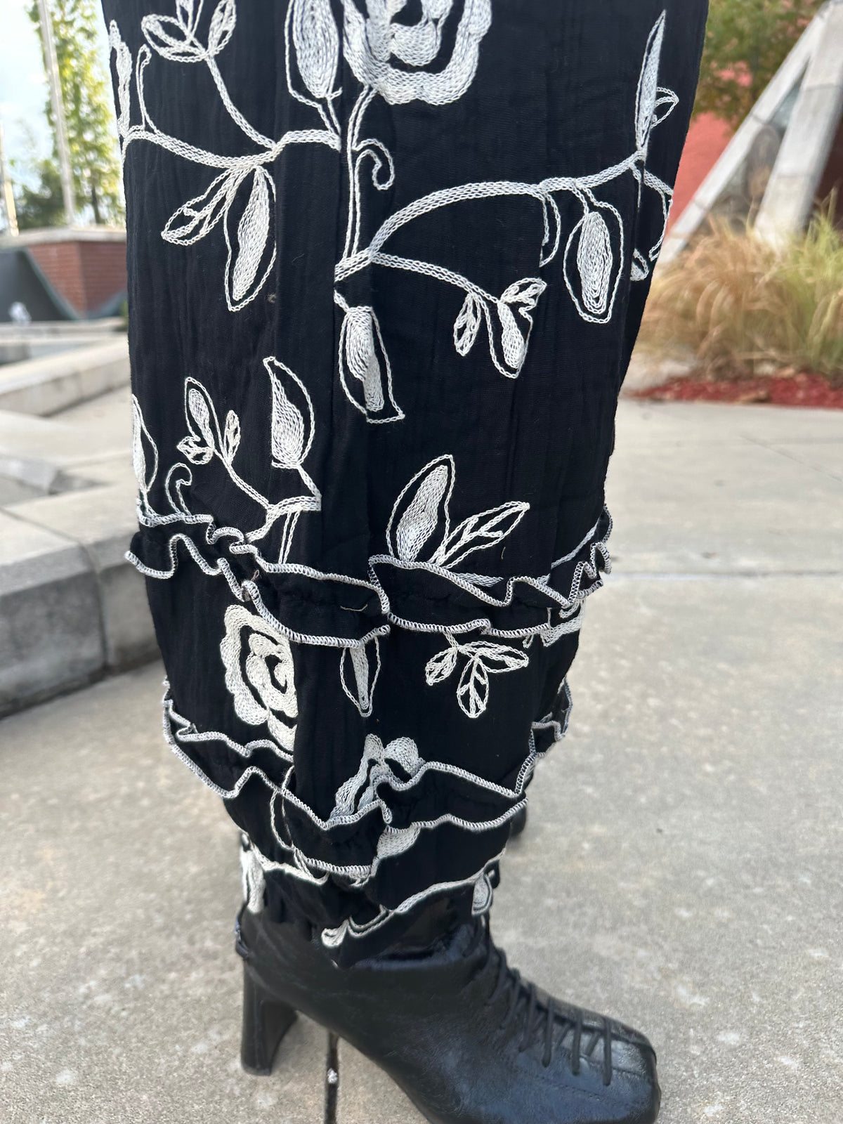 Black pleated pants with white floral embroidery, shown on a model wearing a black lace-up ankle boot in an outdoor setting.