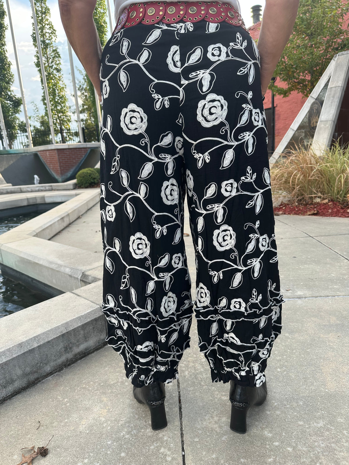 Black wide-leg harem pants with a white floral vine pattern, shown from the back on a model outdoors on a concrete walkway.