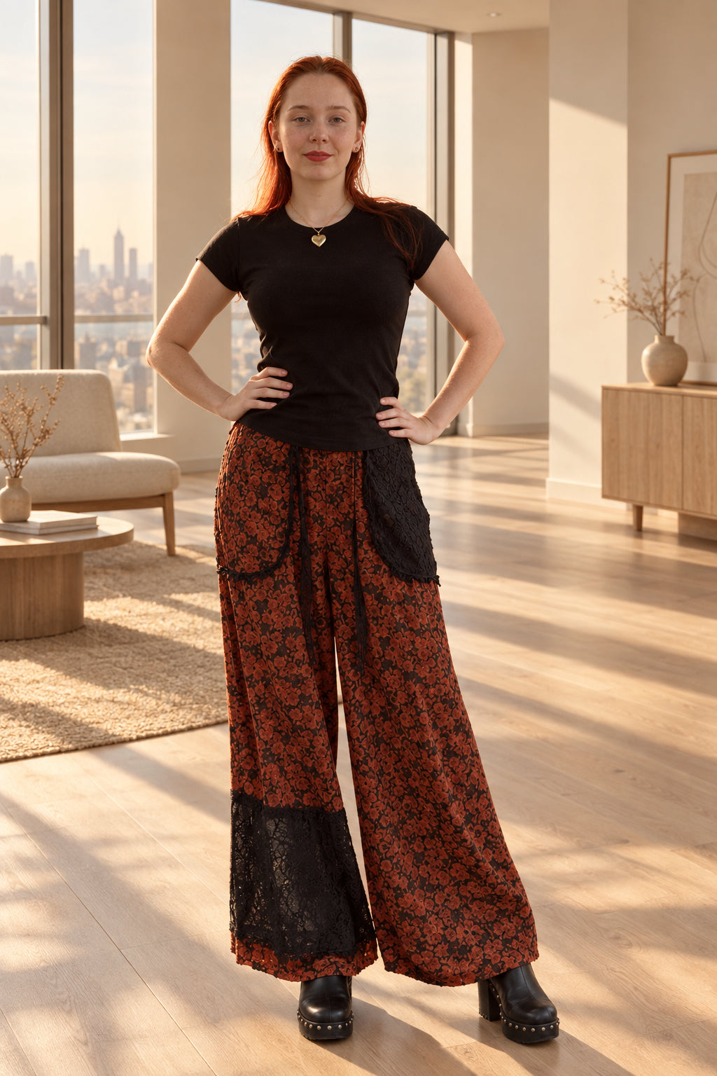 A woman with red hair stands in front of a red brick wall, wearing a black short-sleeve top, wide-leg patterned pants in red and black, and black boots.