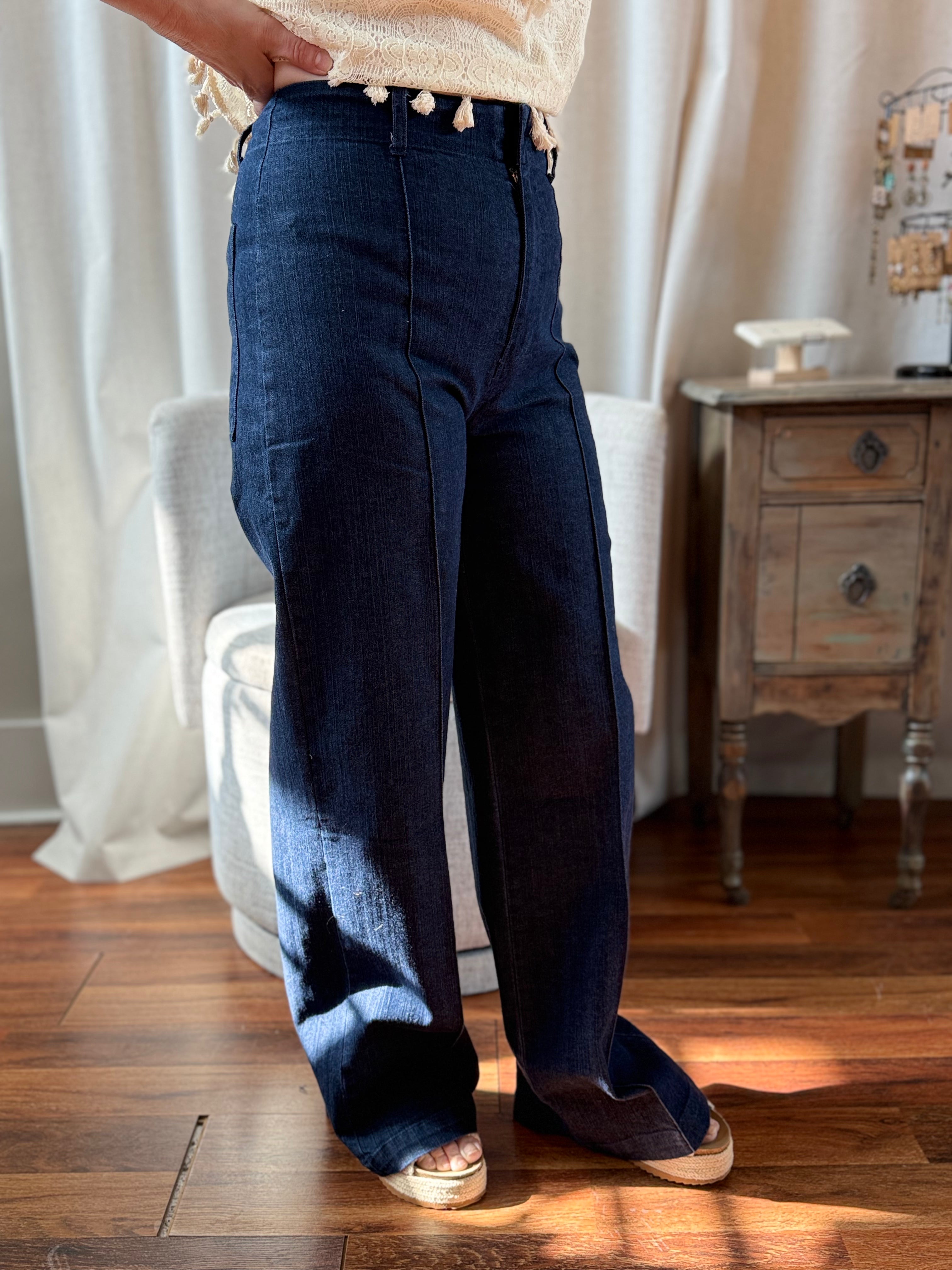 Person wearing blue jeans standing in a room with wooden floor and white curtains.