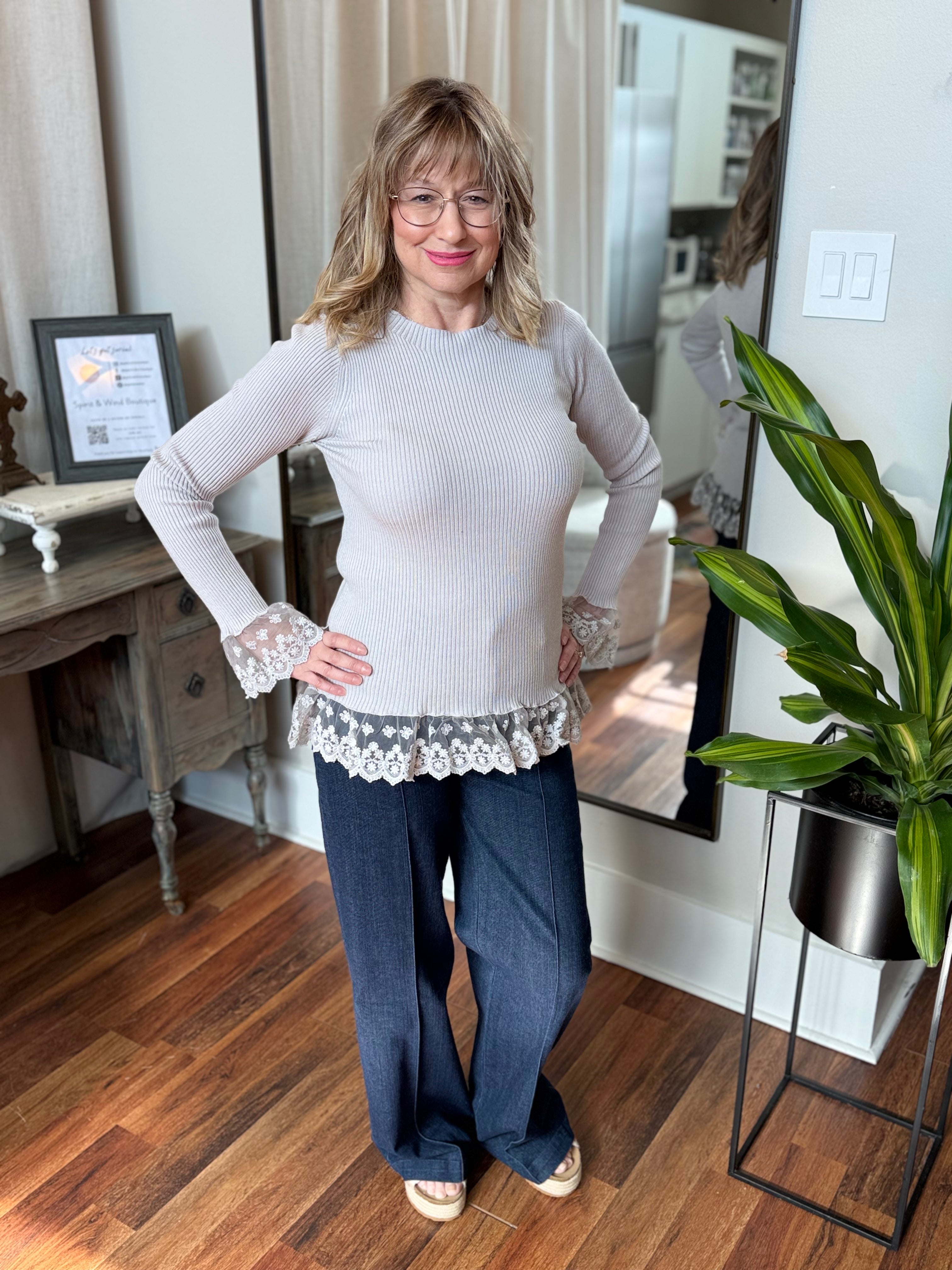 Woman standing in a room with wooden floor and plant, wearing a light gray top and dark blue pants.