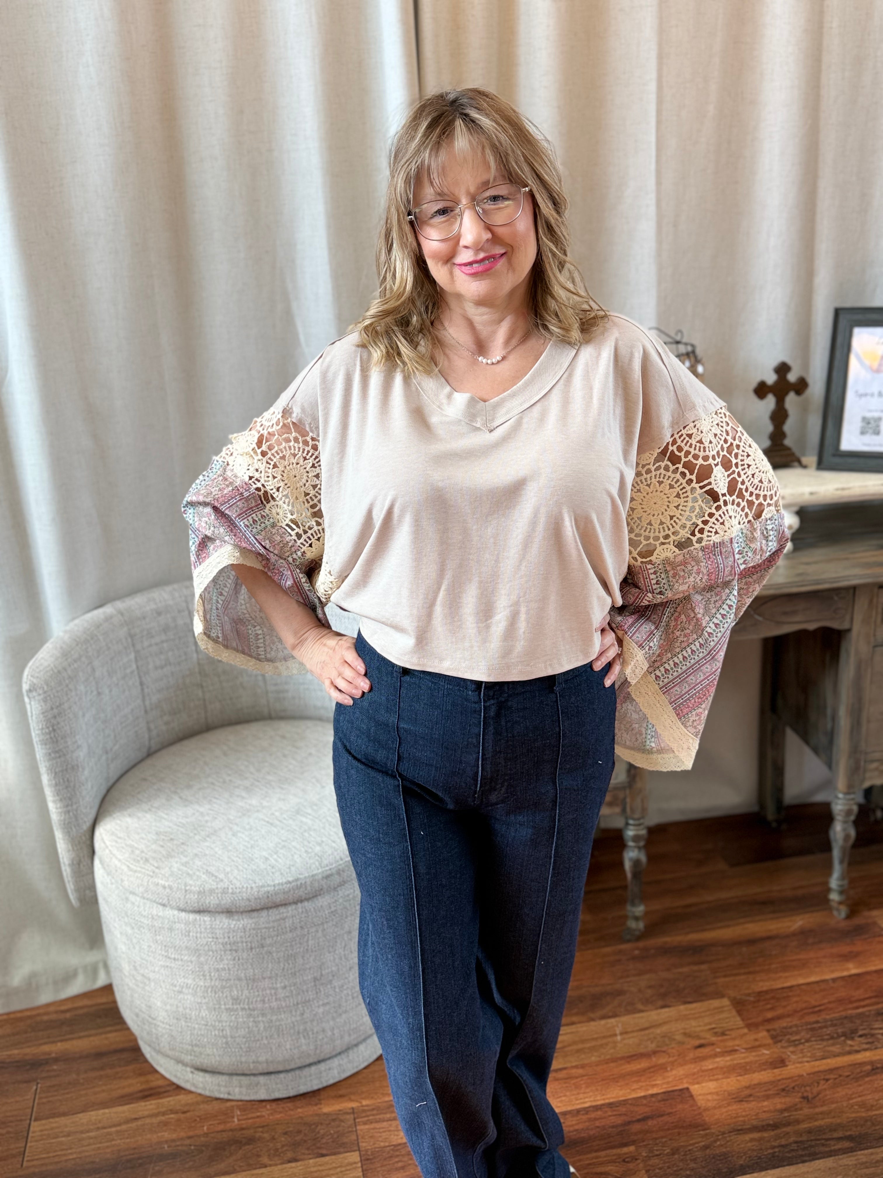 Woman wearing a beige top with floral sleeves and blue jeans standing in a room with a chair and curtain.