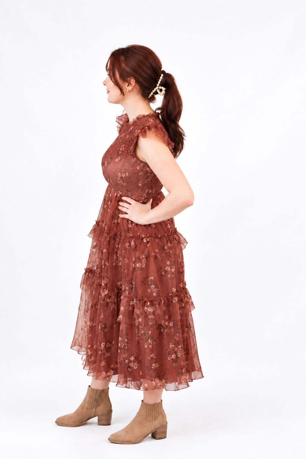 A woman stands in profile wearing a sleeveless rust-brown lace and tulle midi dress with a tiered skirt, paired with beige ankle boots.