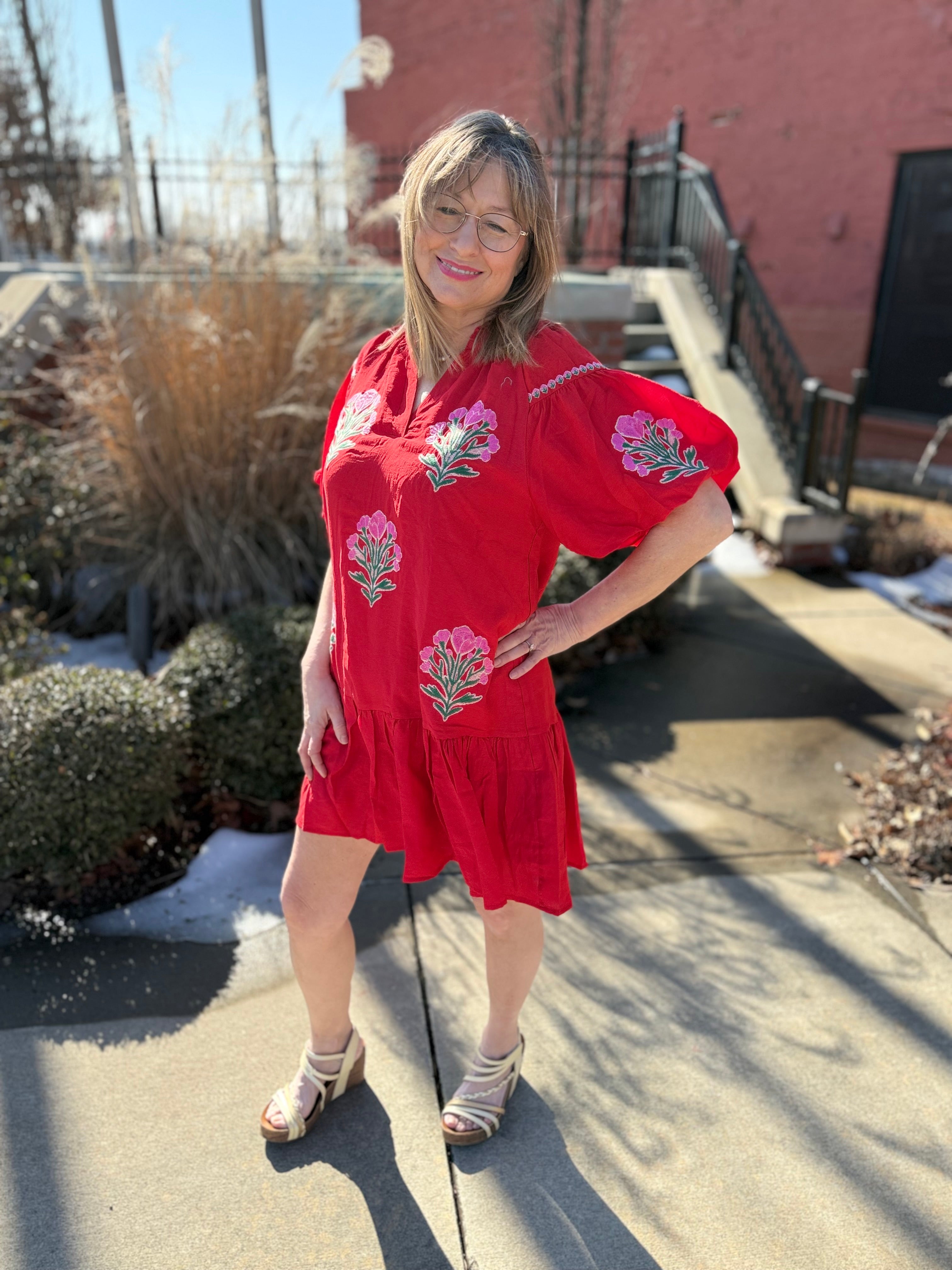 Woman in a red dress with floral embroidery standing outdoors on a sunny day.